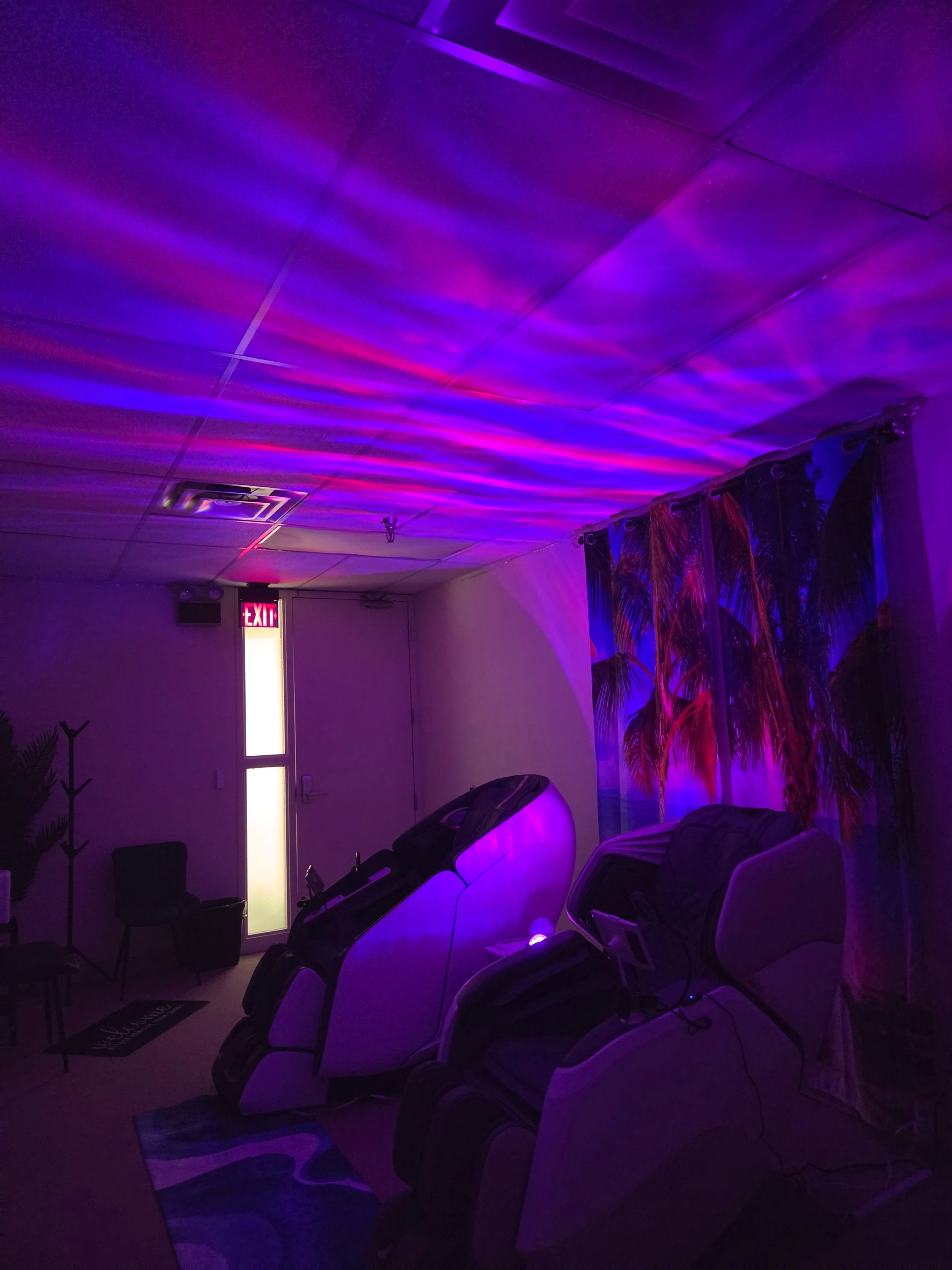 Room with purple and blue LED lighting, massage chairs, a chair, a coat rack, an exit door with an illuminated exit sign, and a curtain with a palm tree pattern.