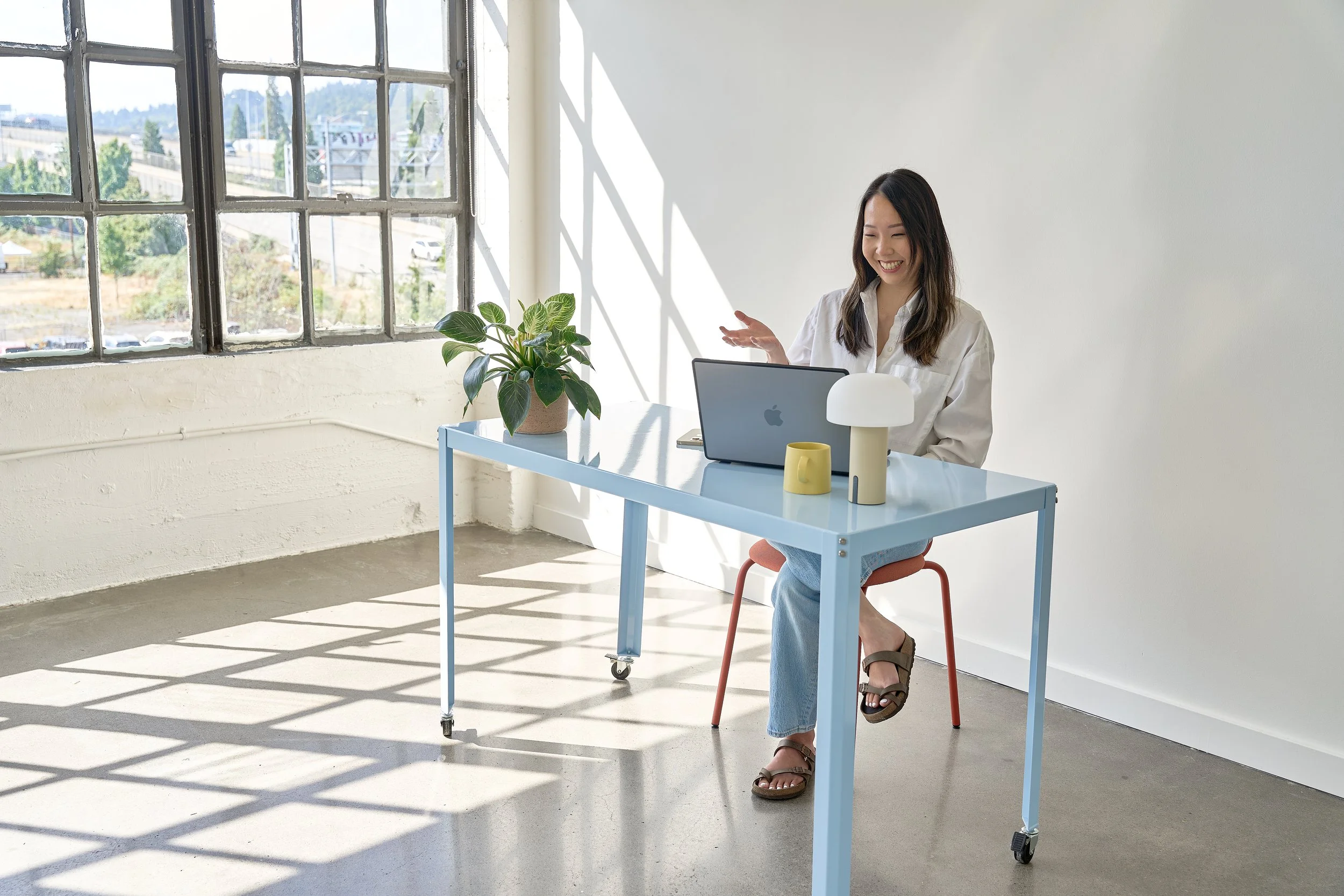 A woman sitting at a light blue desk near a large window with a potted plant, smiling at her laptop.