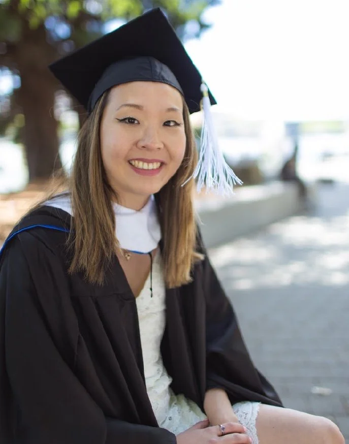 A young woman in a graduation cap and gown sitting outdoors, smiling at the camera.