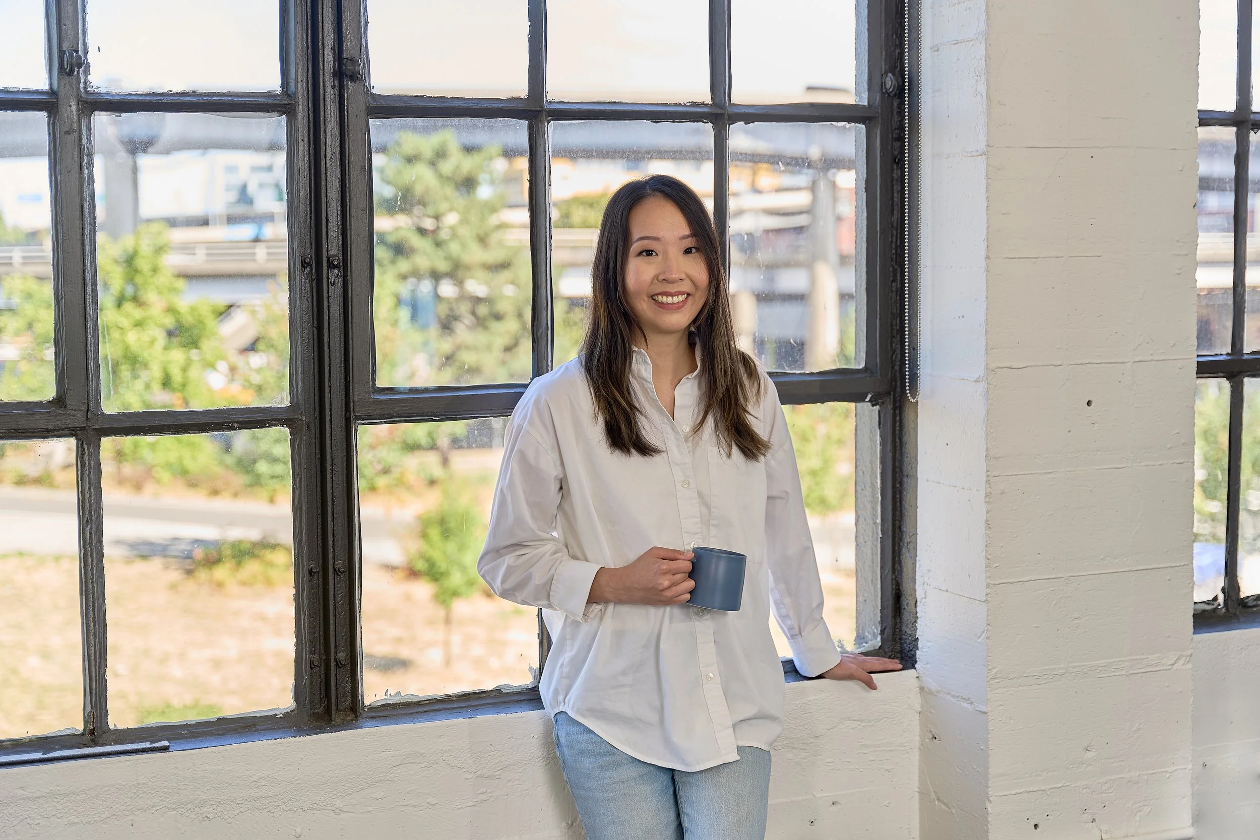 Business coach for therapists wearing a white shirt, standing by a large industrial window with a city view, holding a blue mug, smiling.