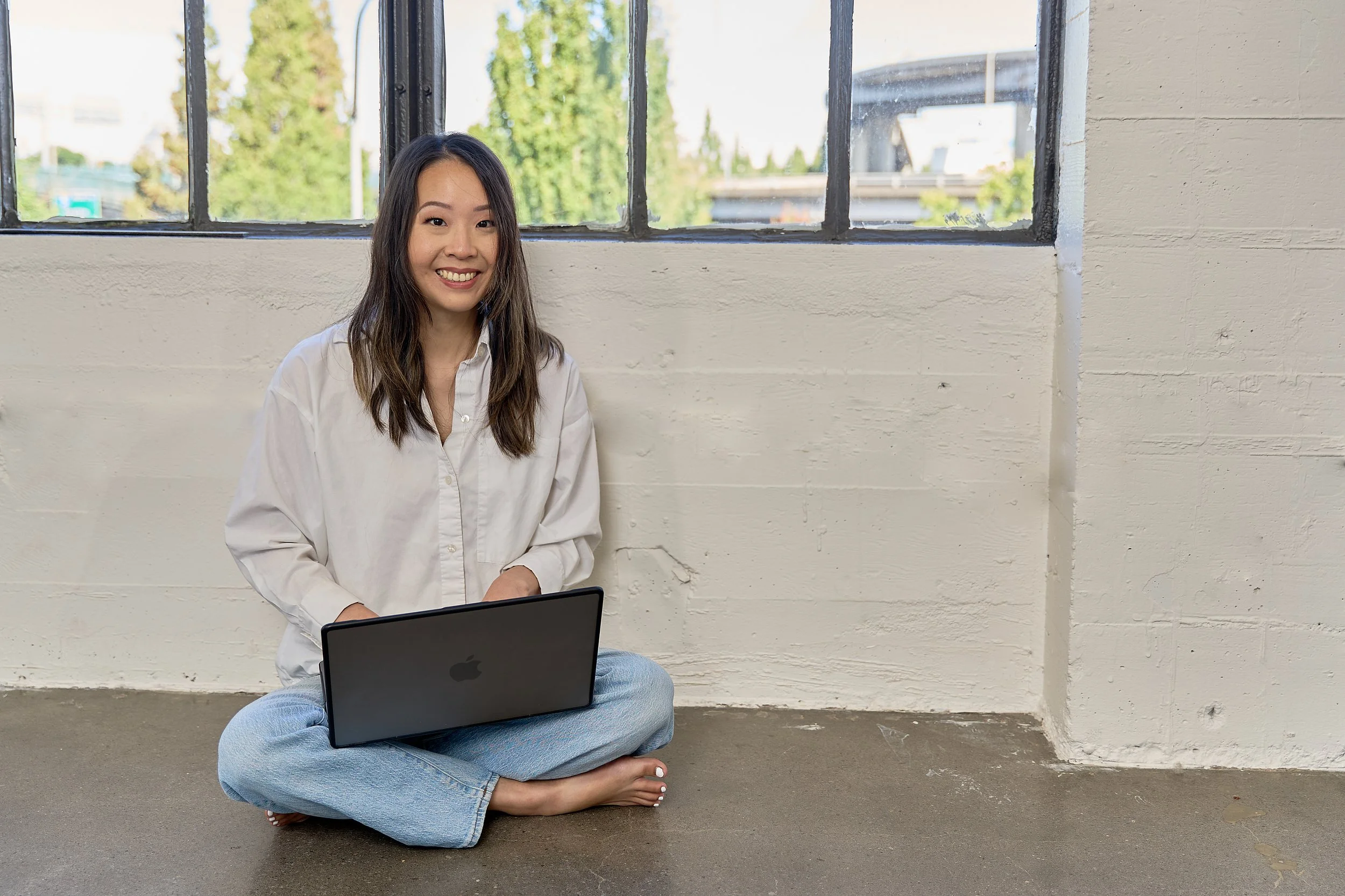 A woman with long dark hair, wearing a white button-up shirt and blue jeans, is sitting cross-legged on the floor in front of a wall with a large window. She is smiling and looking at the camera with a laptop in her lap.
