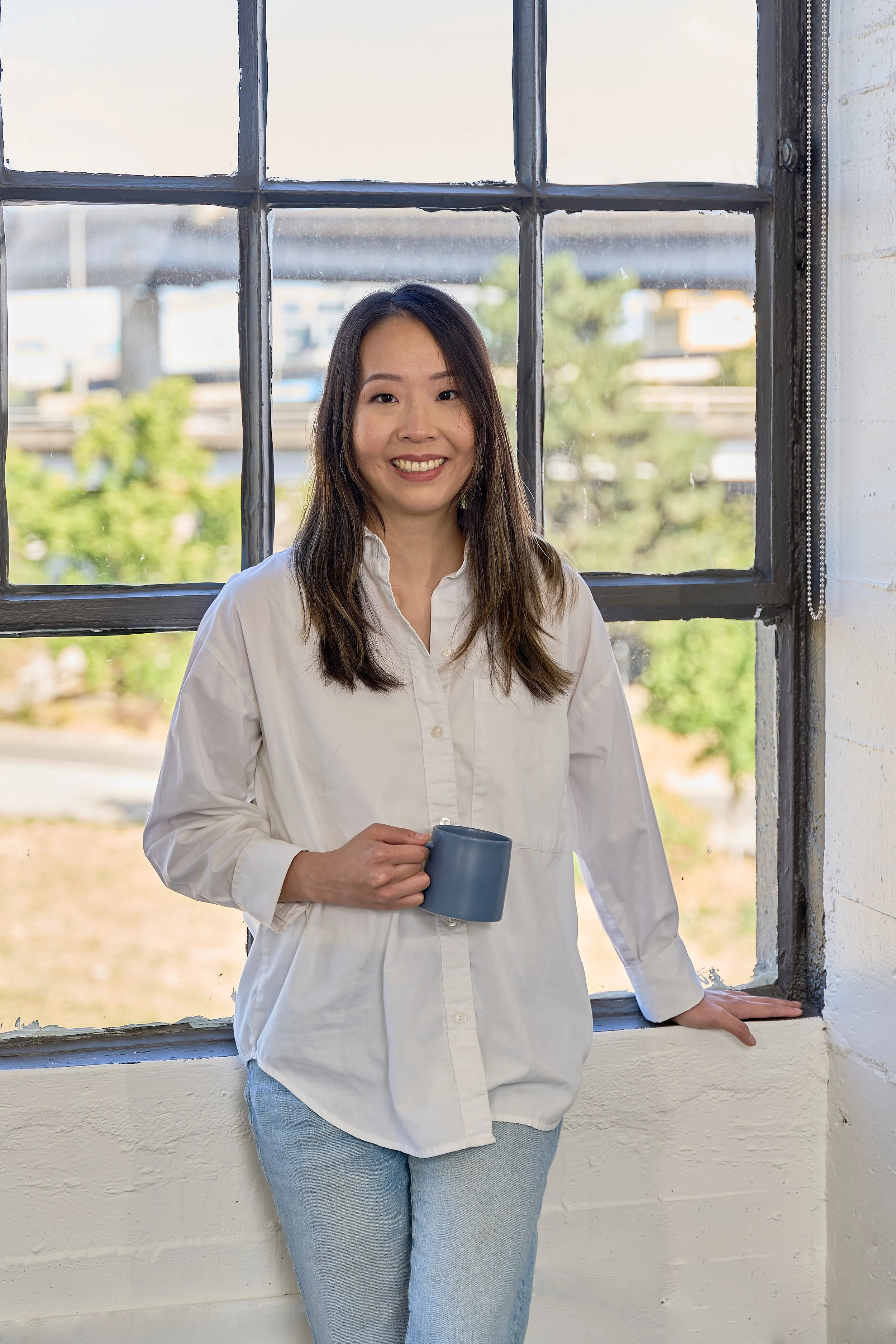 Business coach for therapists with long dark hair, wearing a white button-up shirt and light blue jeans, holding a gray mug, standing inside near a large window in a bright room.