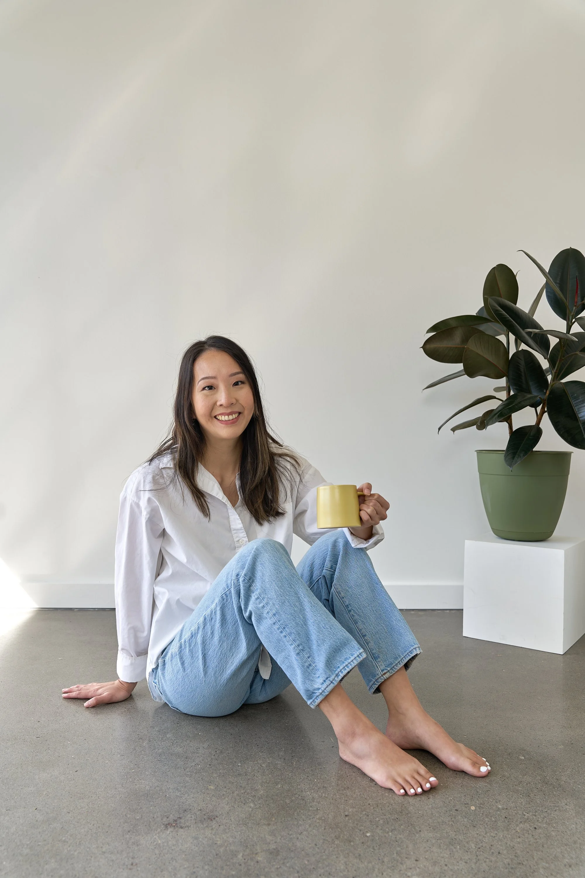A young woman with long dark hair, sitting on a gray floor against a plain light-colored wall. She is smiling, wearing a white shirt and light blue jeans, holding a yellow mug. To her right, there is a large potted plant with dark green leaves in a green pot placed on a white cube.