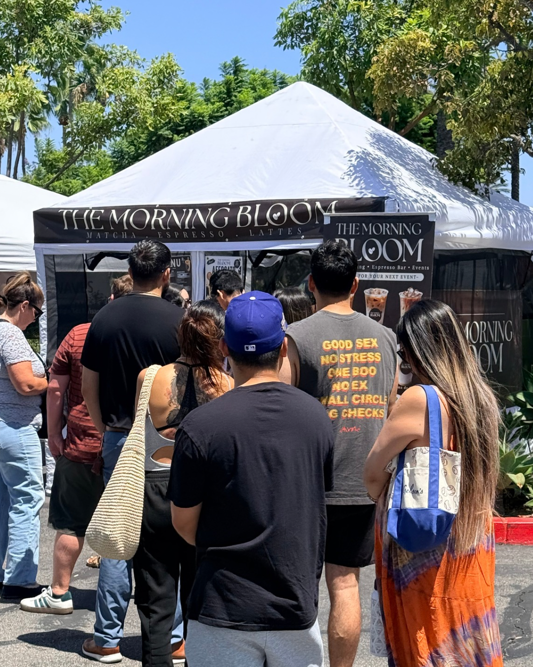 People standing in line at a food truck called 'The Morning Bloom' with signs advertising matcha, espresso, and lattes on a sunny day.