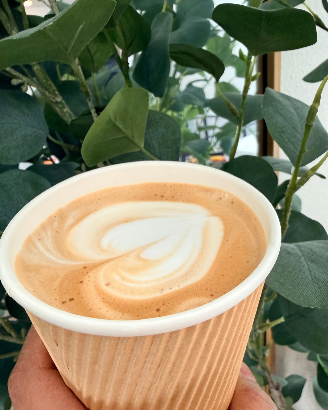 A cup of coffee with latte art in a brown paper cup, held in front of green leafy plants.