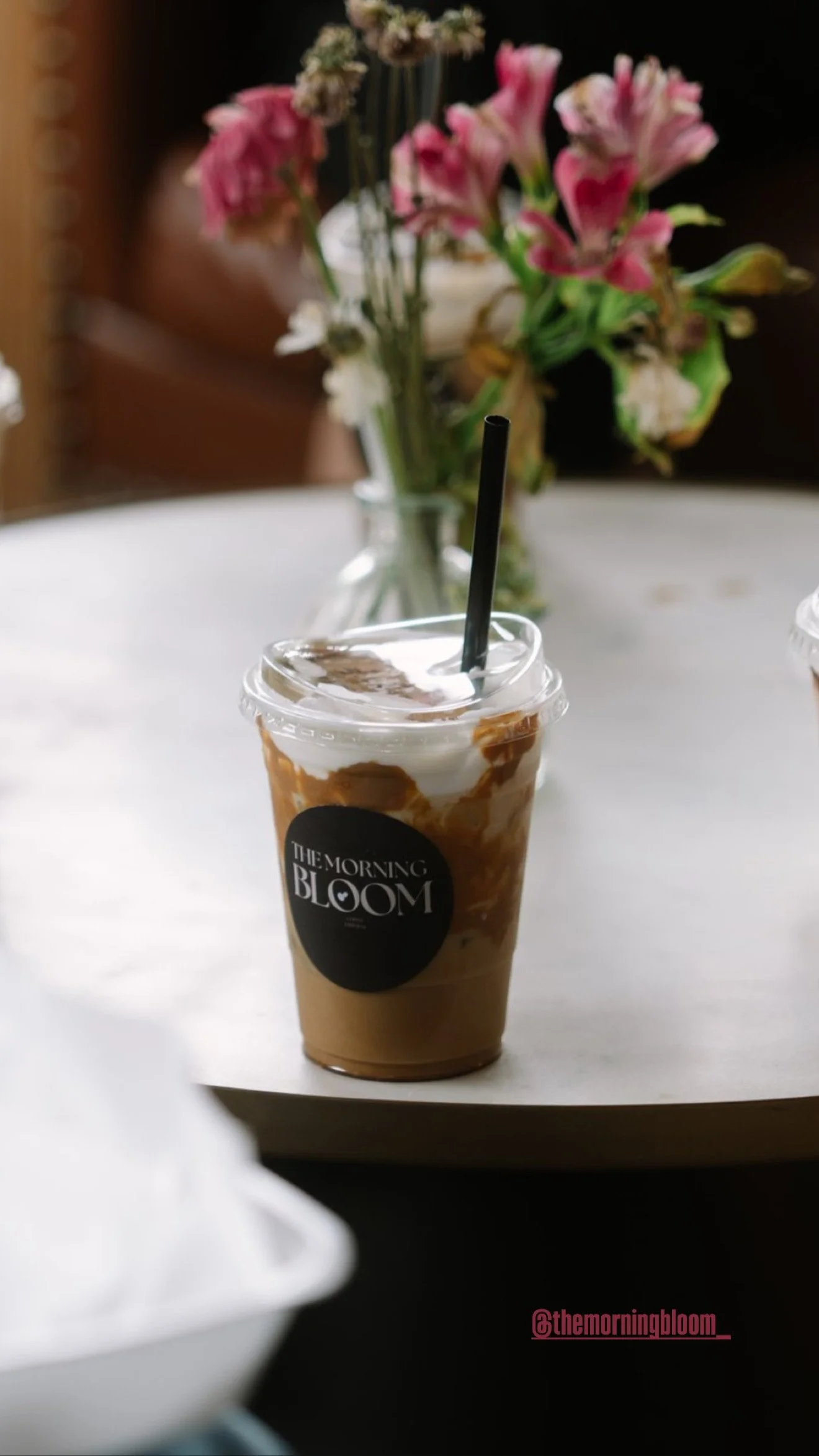 A cold coffee drink in a clear plastic cup with a black straw, labeled 'The Morning Bloom,' on a white table. In the background, there's a vase with pink and white flowers.