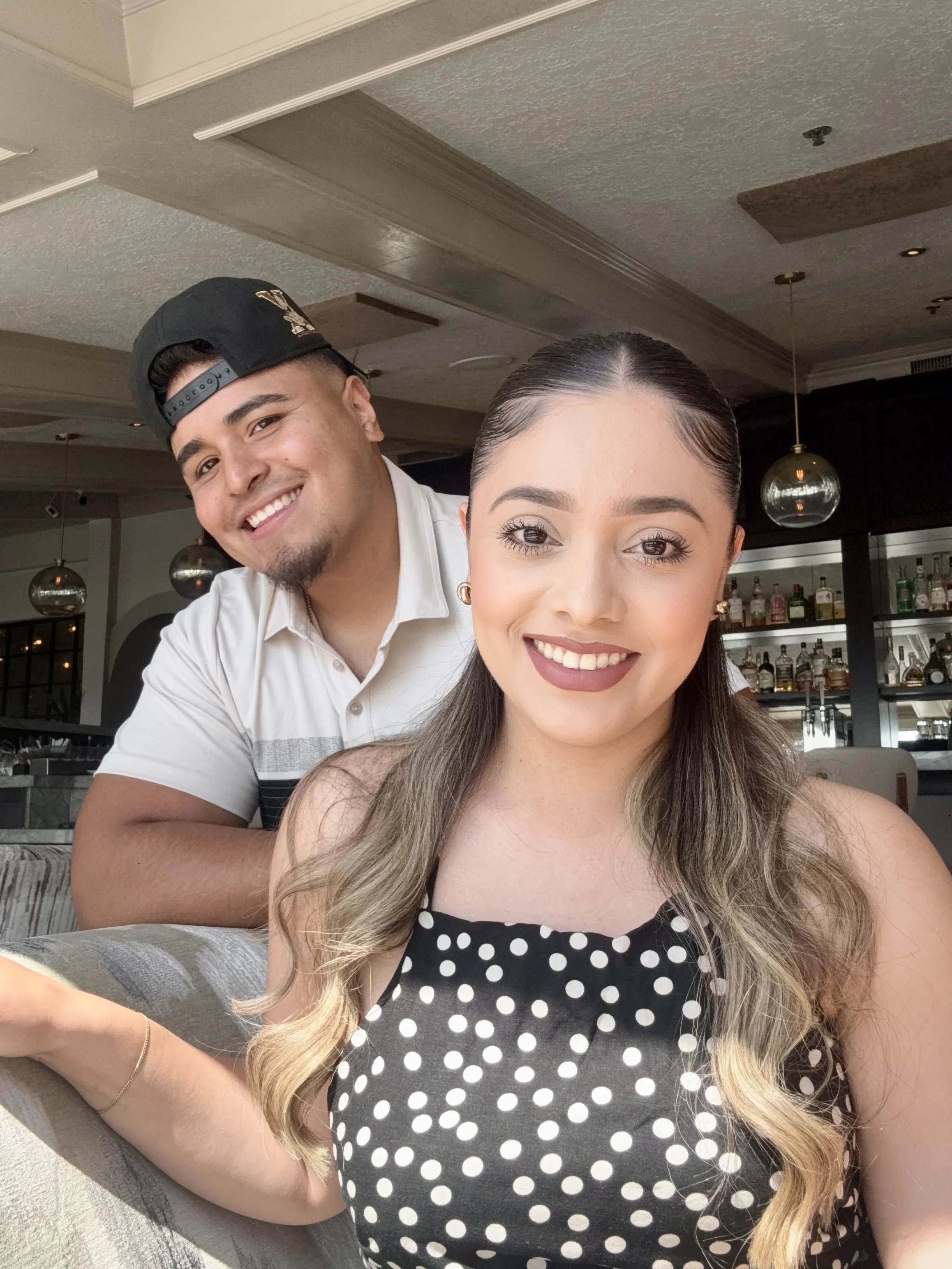 A smiling woman with long hair, wearing a polka dot dress, sitting at a table in a restaurant. Behind her, a man with a black cap and a white shirt is leaning in, smiling.