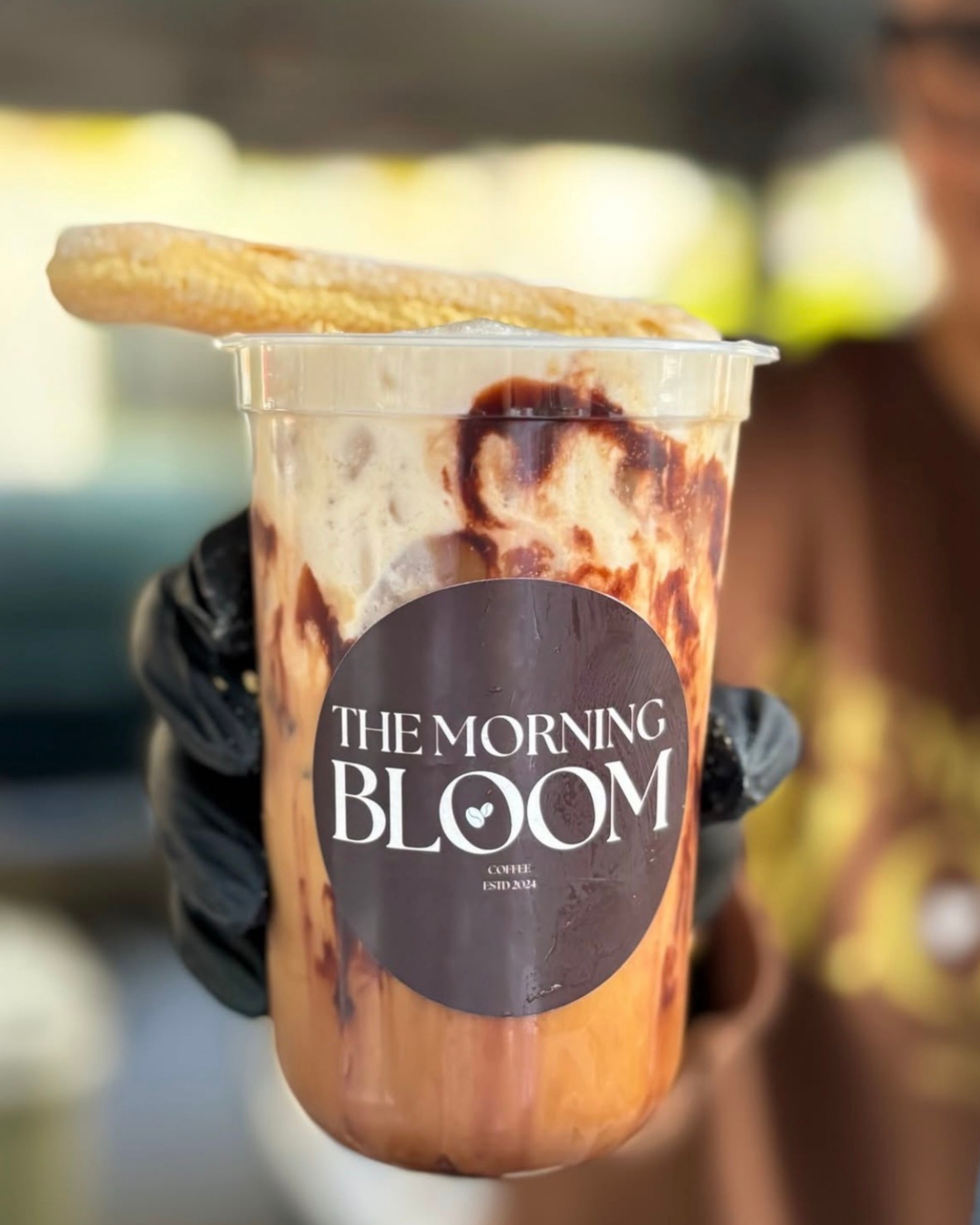Close-up of a iced coffee drink in a plastic cup with a black sleeve and a sticker that reads 'THE MORNING BLOOM', with a cookie on top of the cup, with a blurred background.