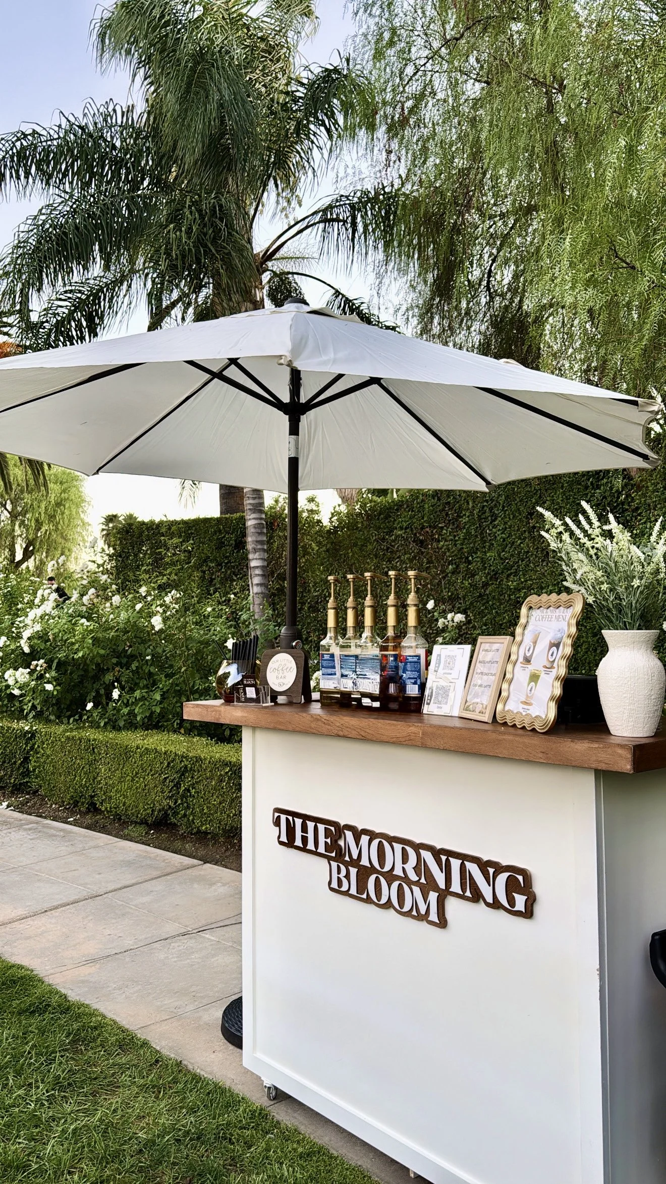 An outdoor coffee stand with a white umbrella, bottles of flavored syrup, and a framed menu, with lush green trees and bushes in the background.