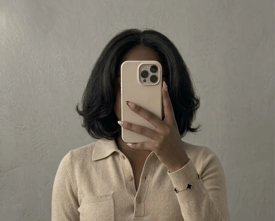 A woman with shoulder-length dark hair taking a mirror selfie with a beige smartphone, standing against a plain textured wall, wearing a beige collared shirt.