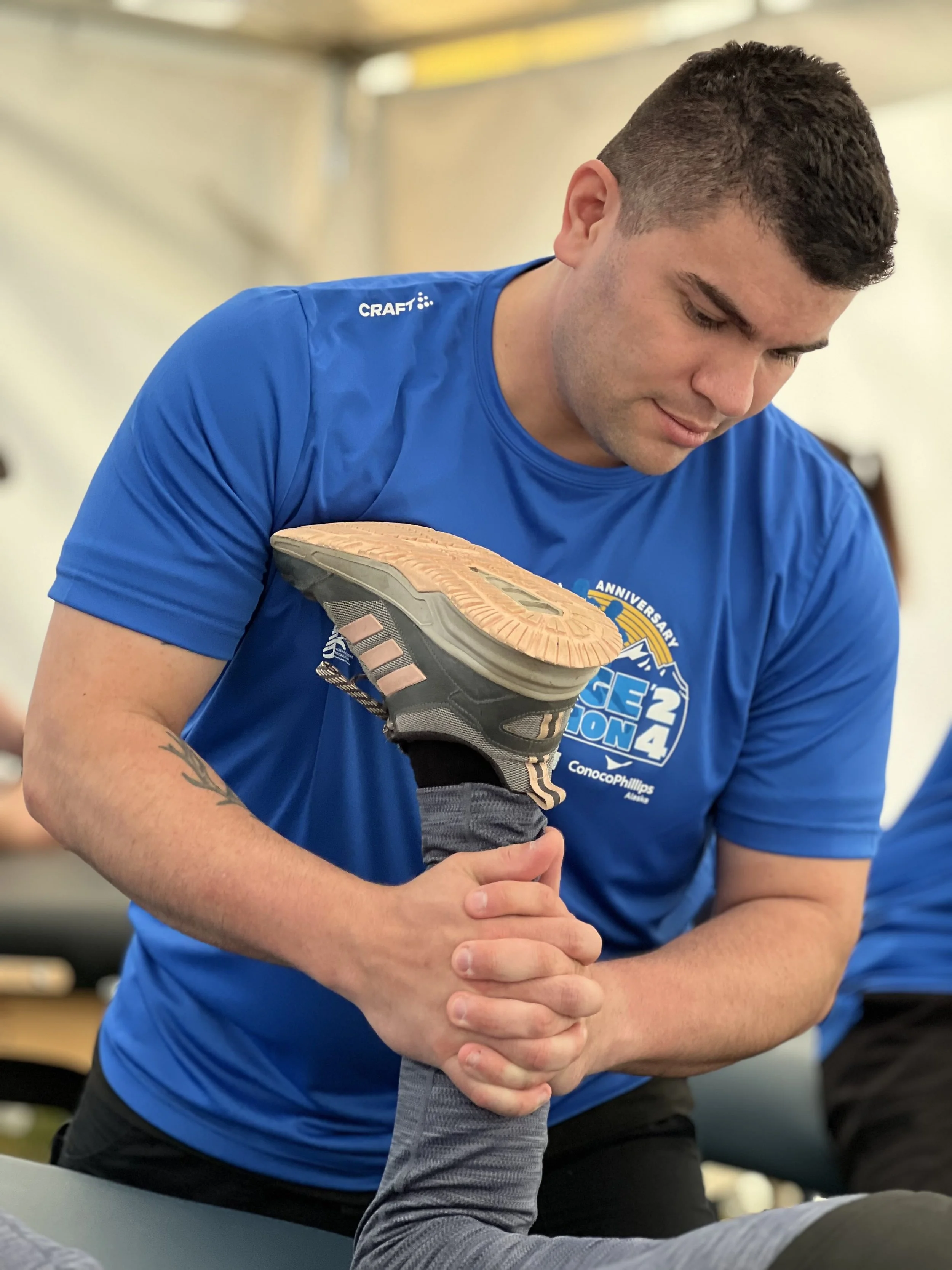 A man in a blue shirt helping another person stretch their leg during an exercise or physical therapy session.