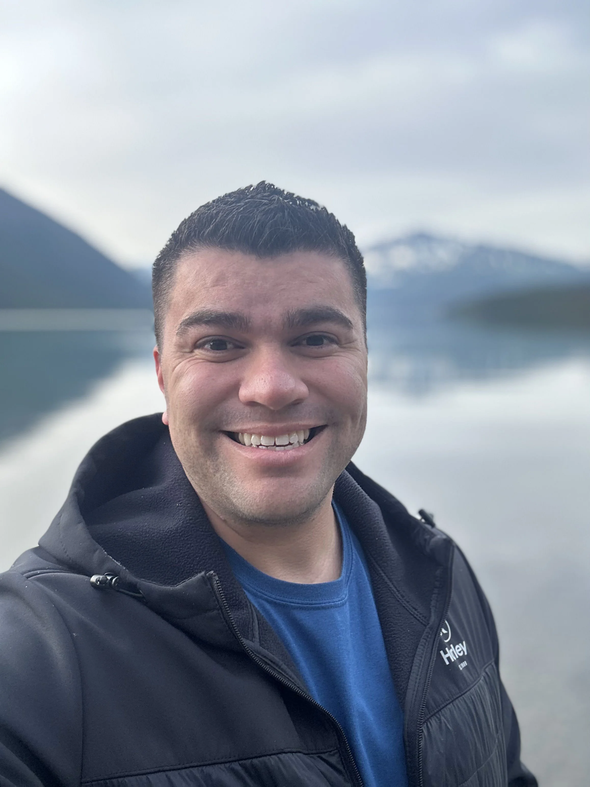 A smiling man in a black jacket and blue shirt taking a selfie outdoors with mountains and water in the background.