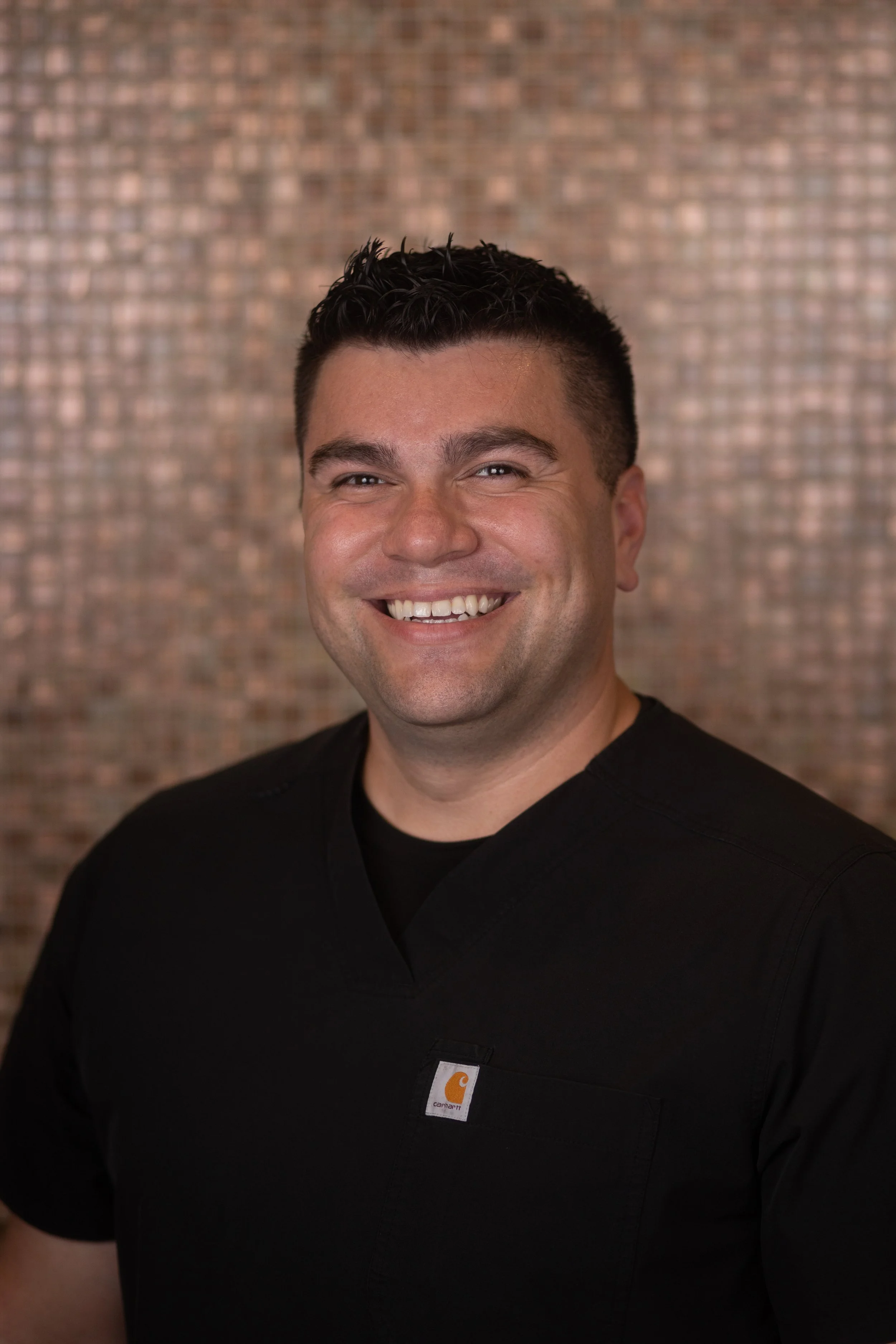 A man smiling, wearing a black Carhartt shirt, standing against a brick wall background.