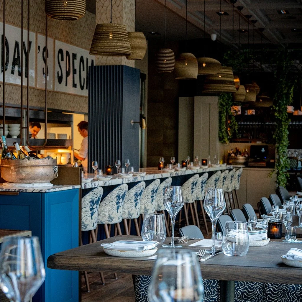 A restaurant interior with a long table, glasses and plates, hanging lamps, and a bar with chairs.