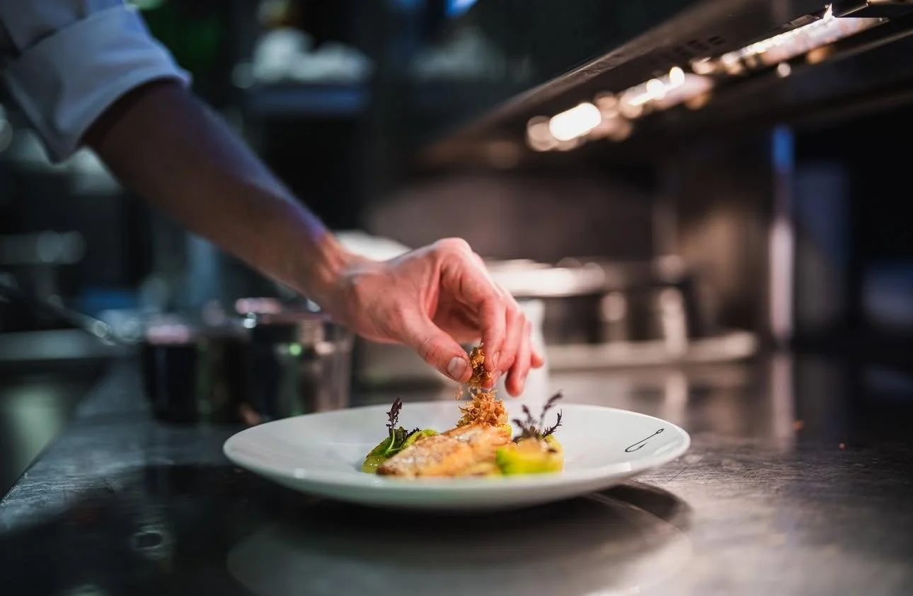 A chef finishing a dish with a garnish on a white plate in a professional kitchen.