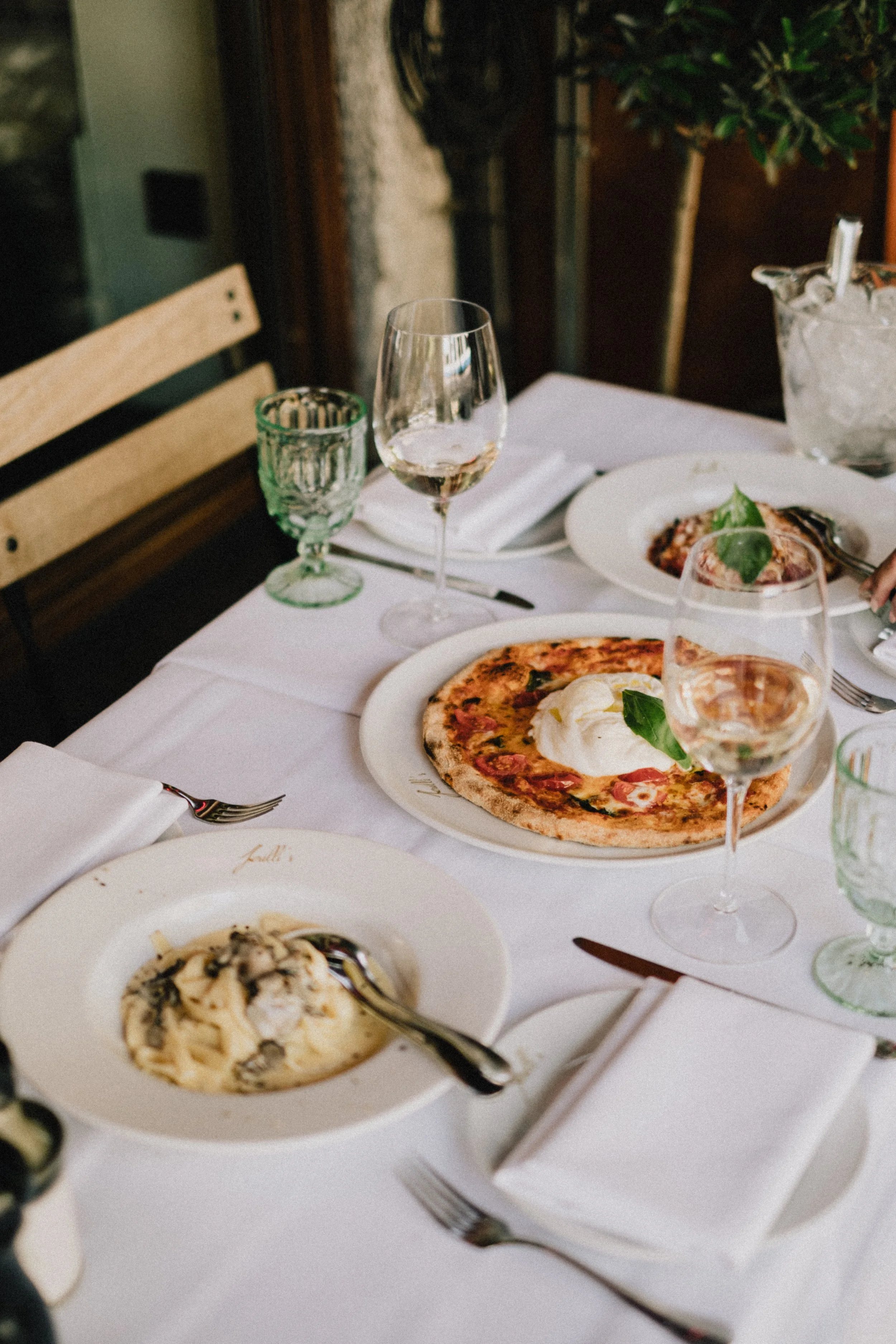 A table with various dishes, including pizza, pasta, and glasses of water and wine, in a restaurant or café.