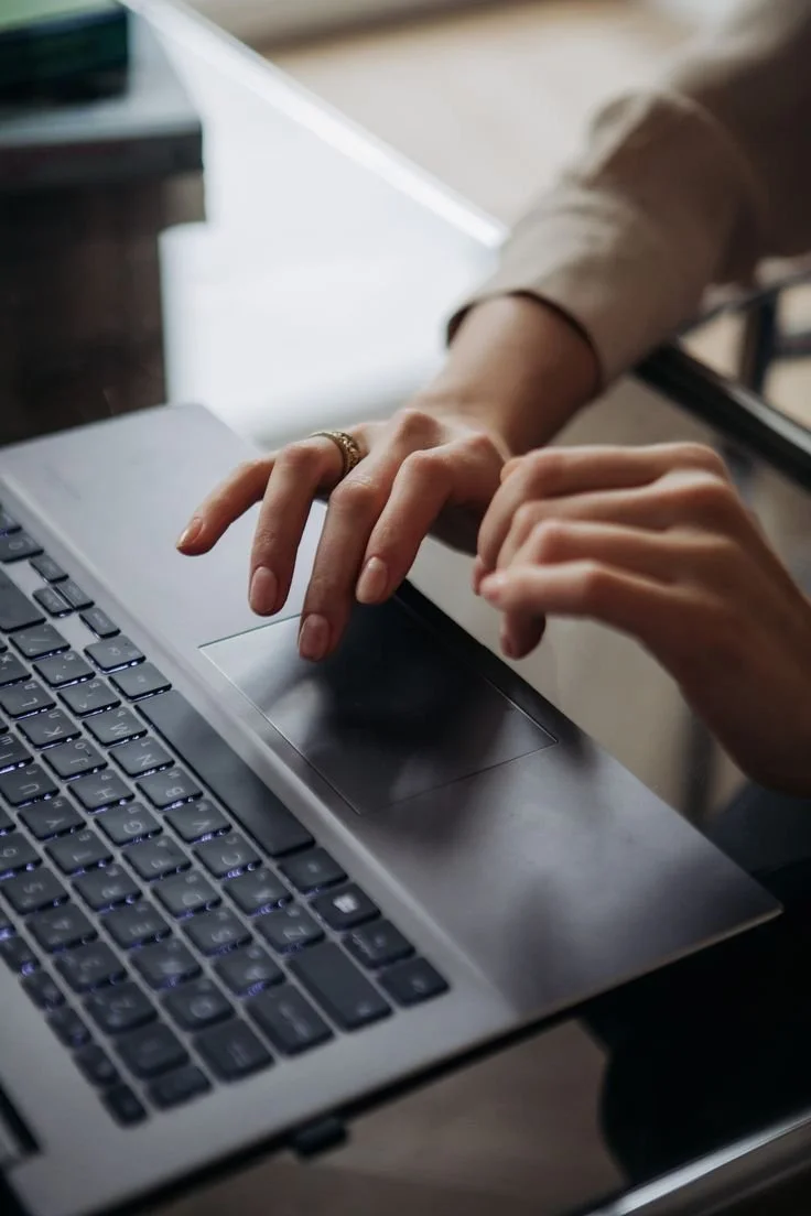 A person is typing on a laptop with a silver case and black keys. The person is wearing a ring and a beige sleeve.