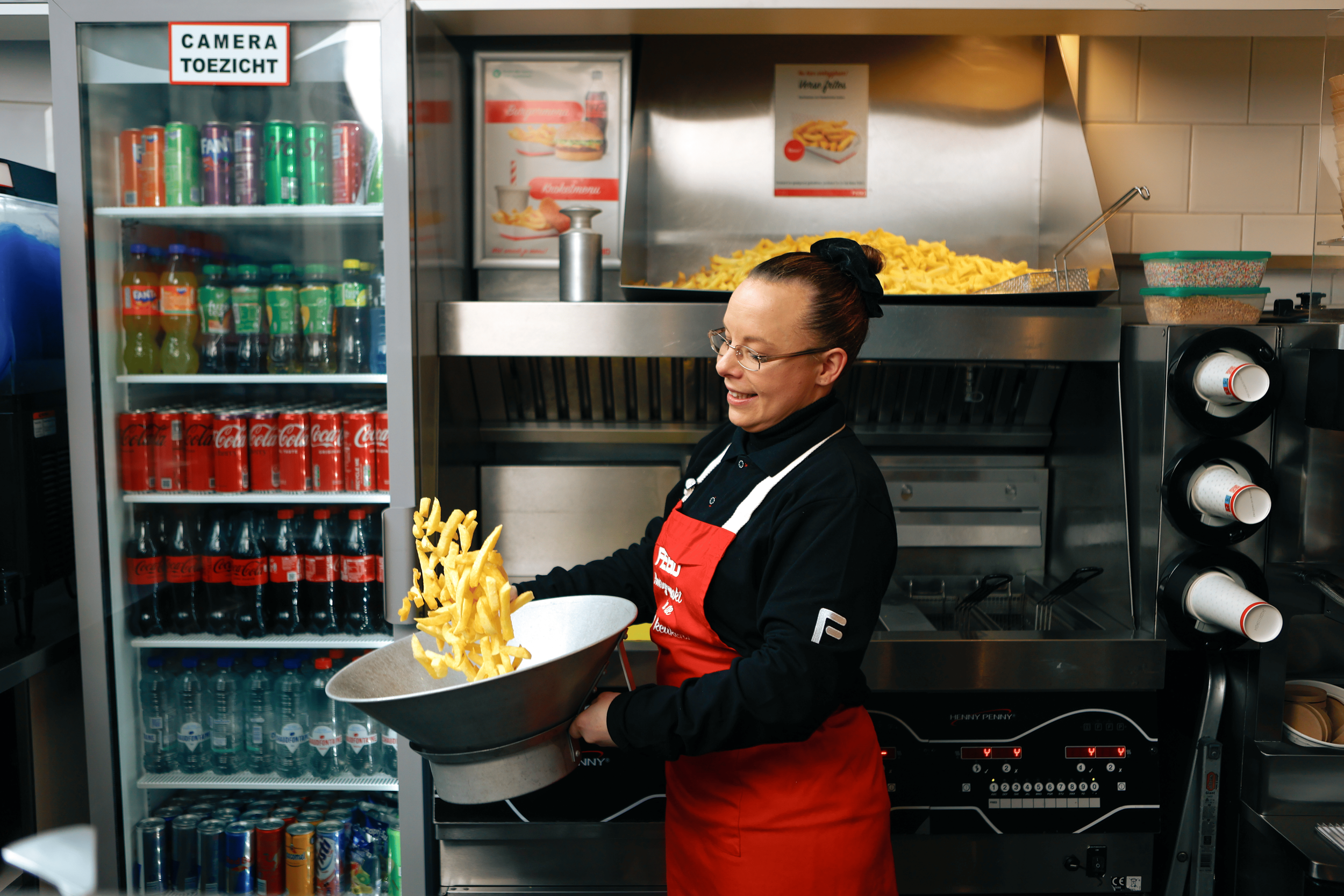 A woman serving french fries at a fast-food restaurant, with a refrigerator full of drinks and frying oil in the background.