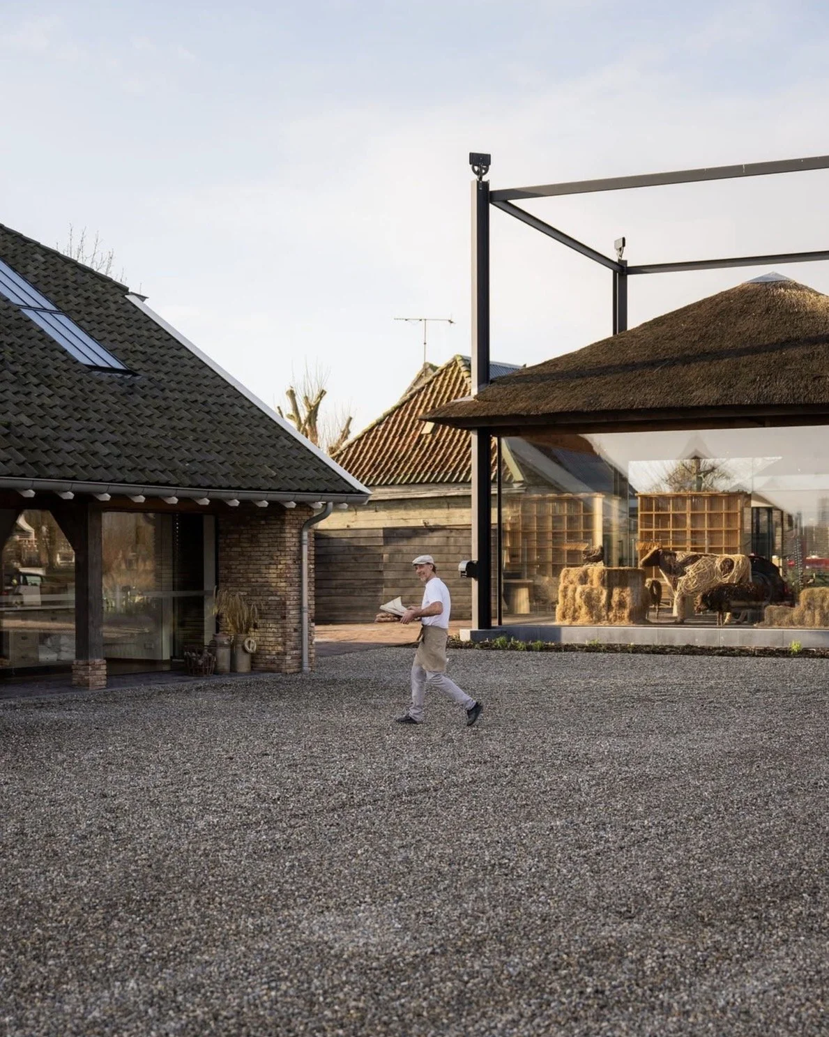 A man walks down a gravel driveway next to modern buildings, one of which has a glass facade filled with bales of hay and decorative props.