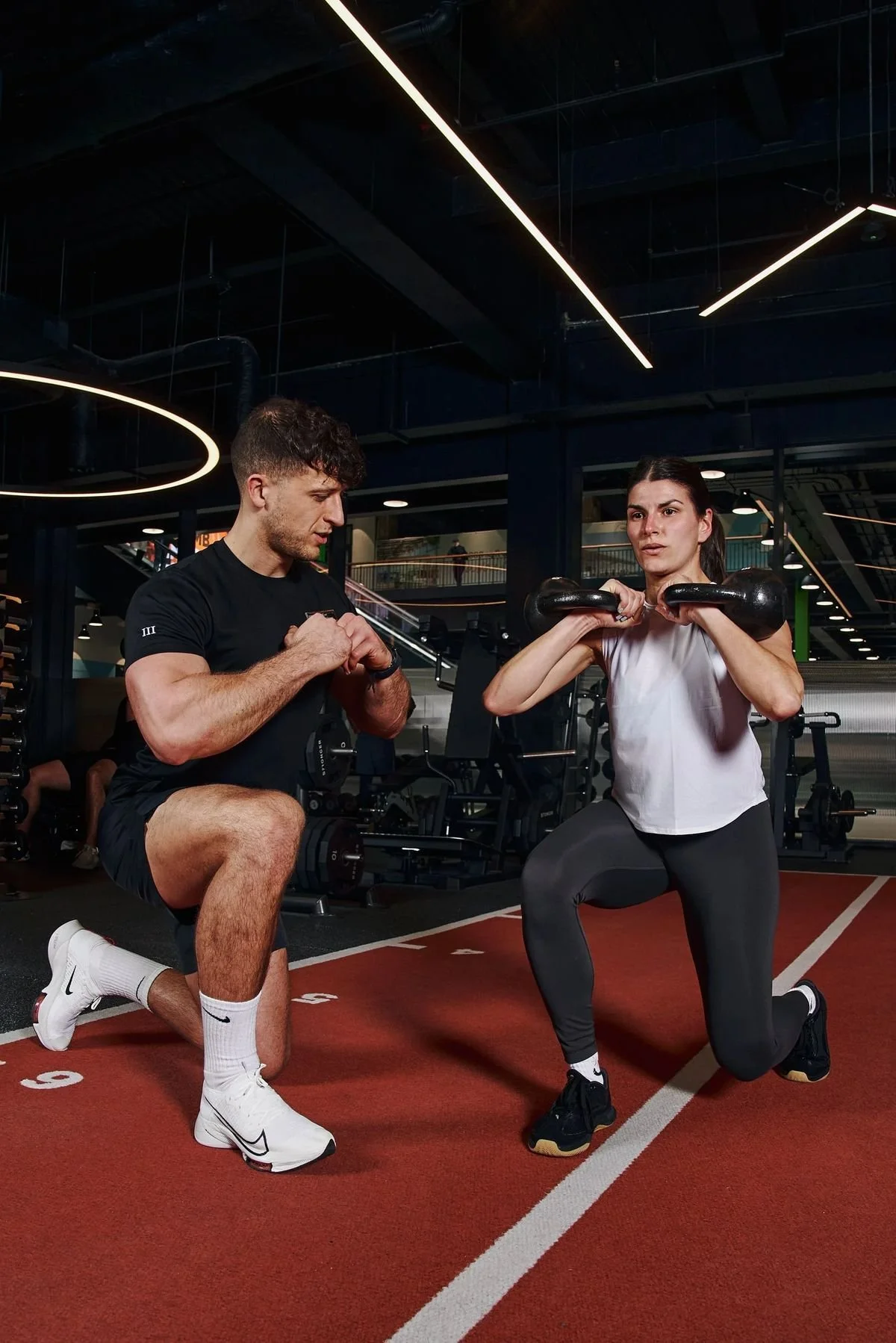 A man and a woman are working out at a gym. The woman is doing squats with a barbell, while the man stands in a crouched position, ready to coach her.