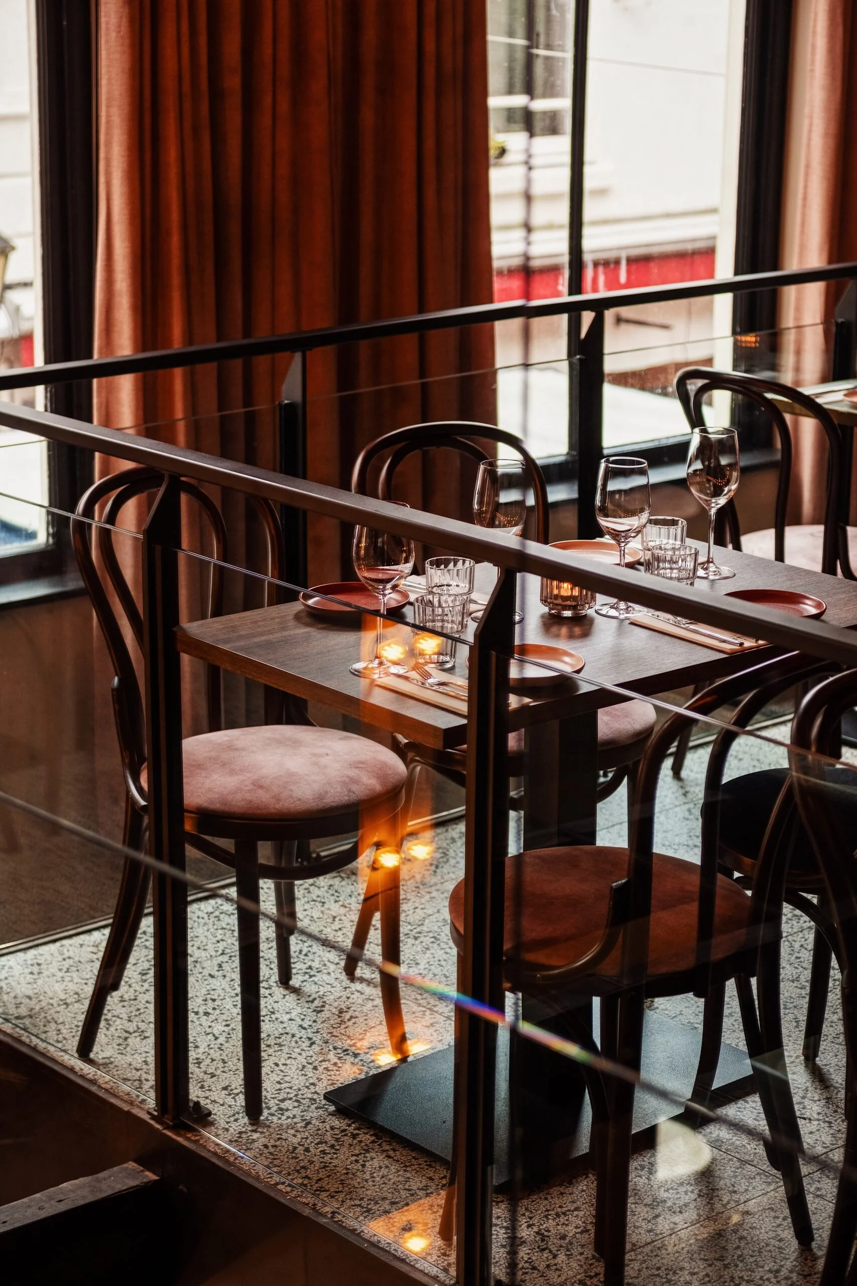 An interior shot of a restaurant with a table set with glassware, plates, silverware, and candles. Large windows with reddish-brown curtains and a view of a balcony and a building outside.