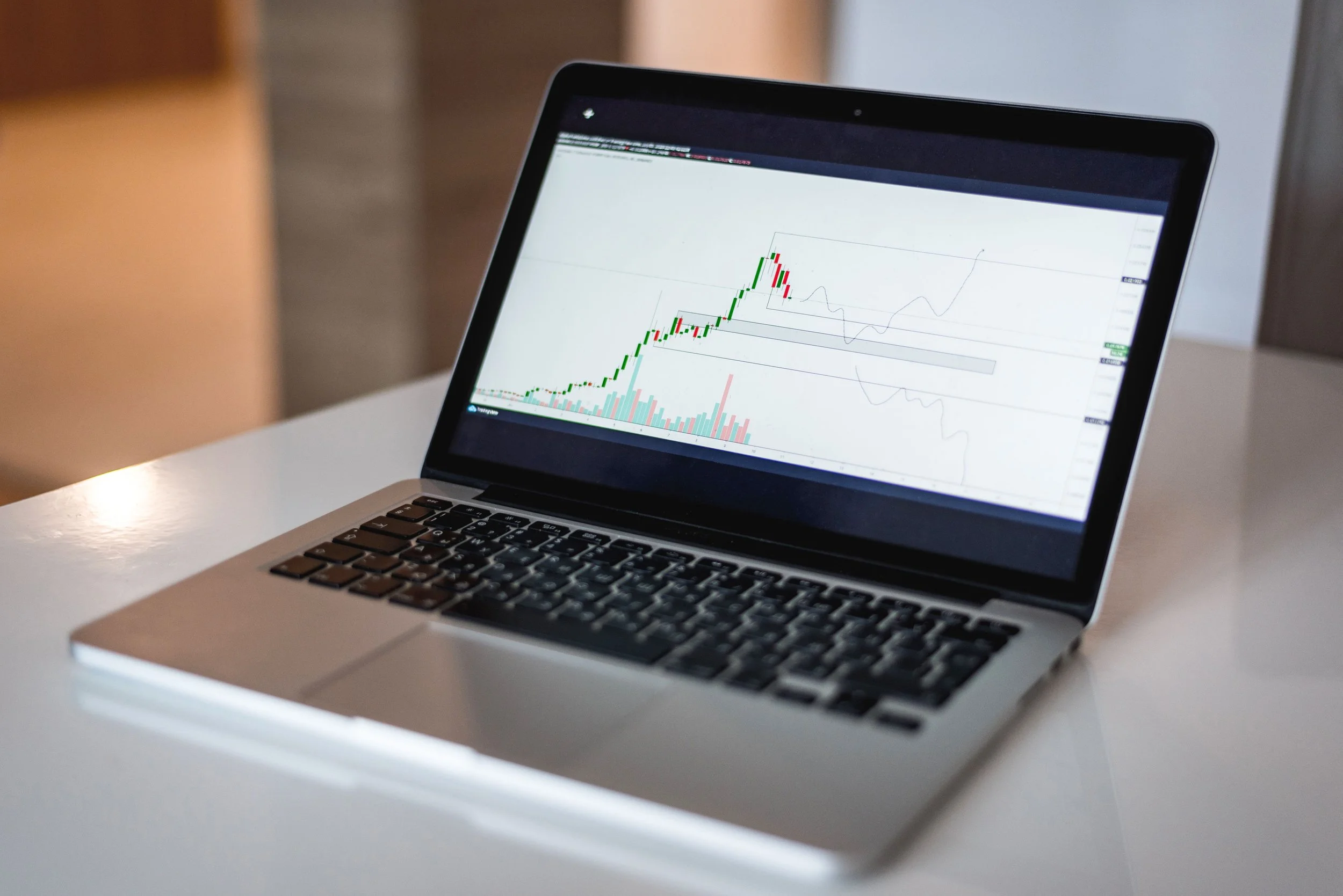The image shows a laptop resting on a white surface. The laptop's screen displays a chart with green and red candlesticks and several line charts, possibly related to stock or cryptocurrency trading.