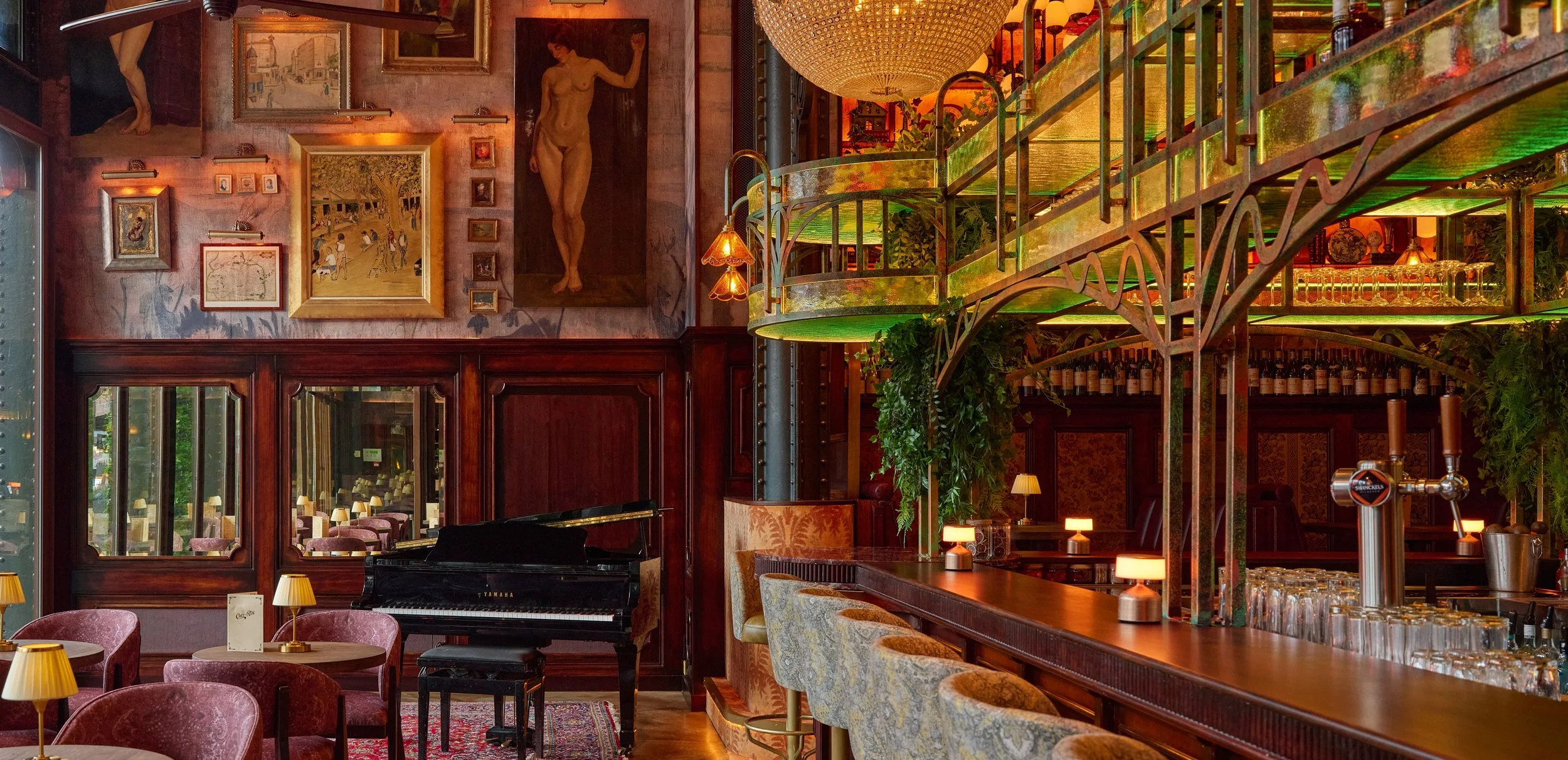 The interior of a restaurant or café featuring rich wooden paneling, artwork on the walls, a piano, and a bar with glasses and taps, illuminated by warm yellow light and decorative lamps.