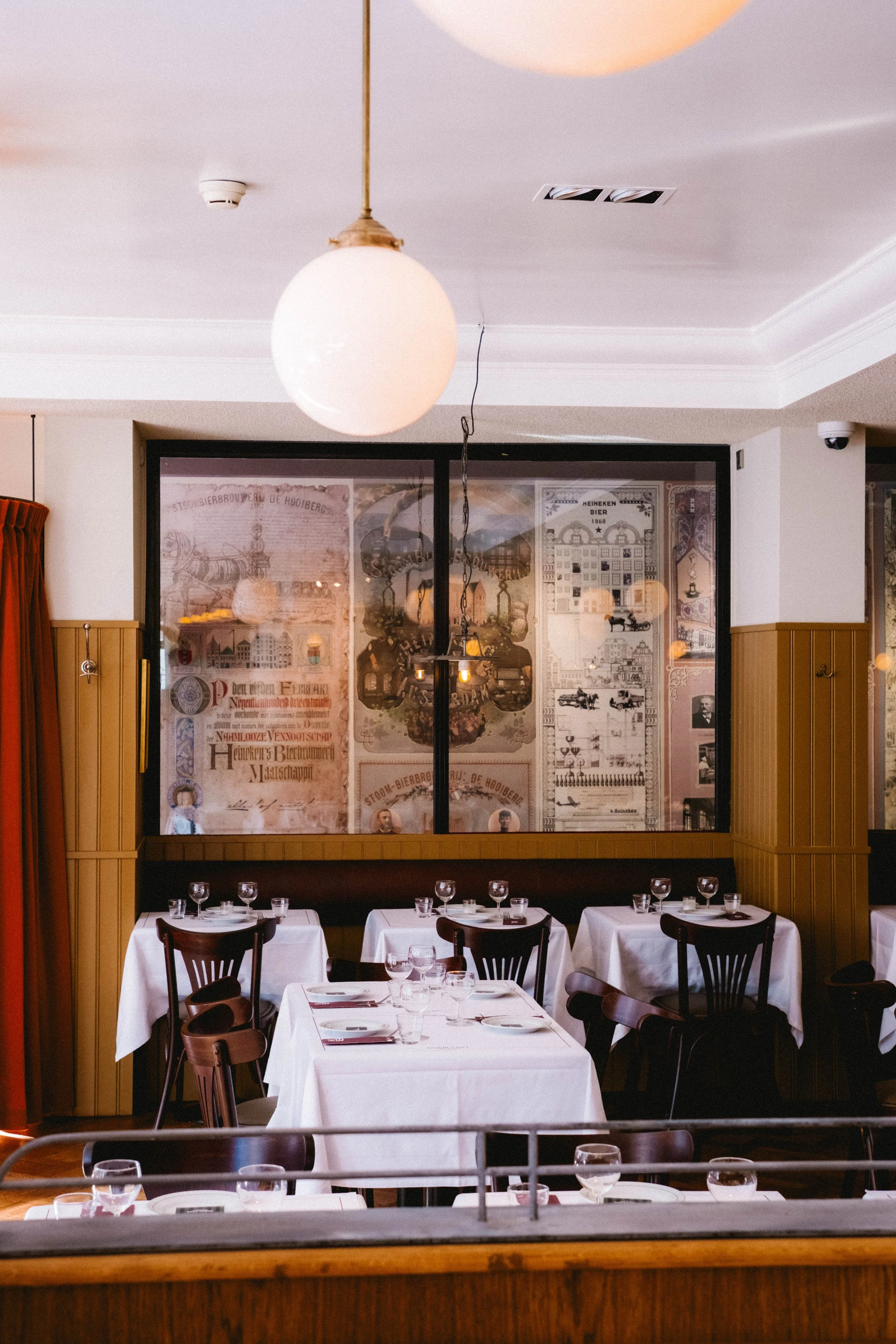 A restaurant with tables set with white tablecloths and black chairs, wall decorations featuring old posters and photos, and a round light fixture hanging from the ceiling.