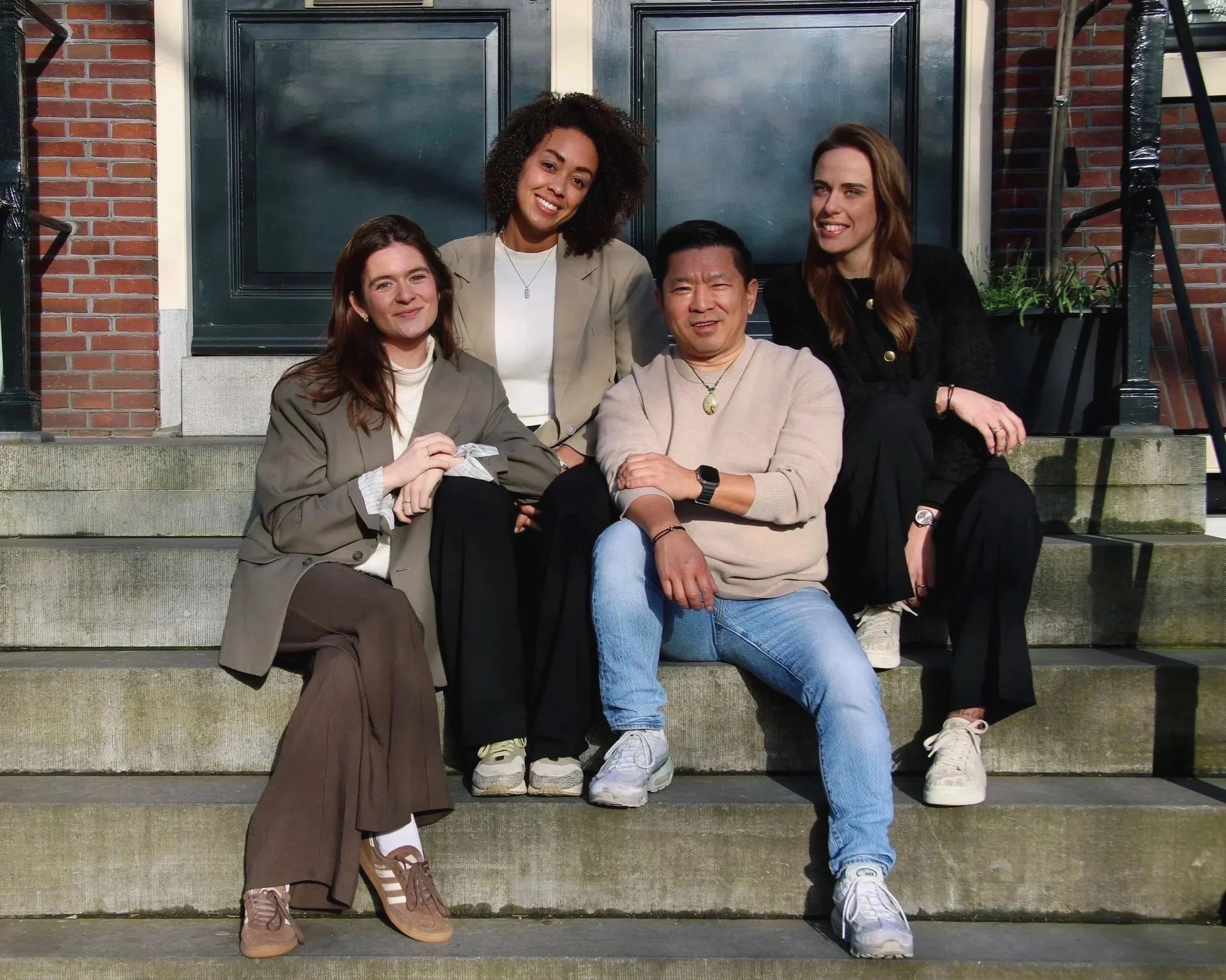 Five people are sitting and standing on the steps in front of a house with brick walls, smiling and posing for the photo.