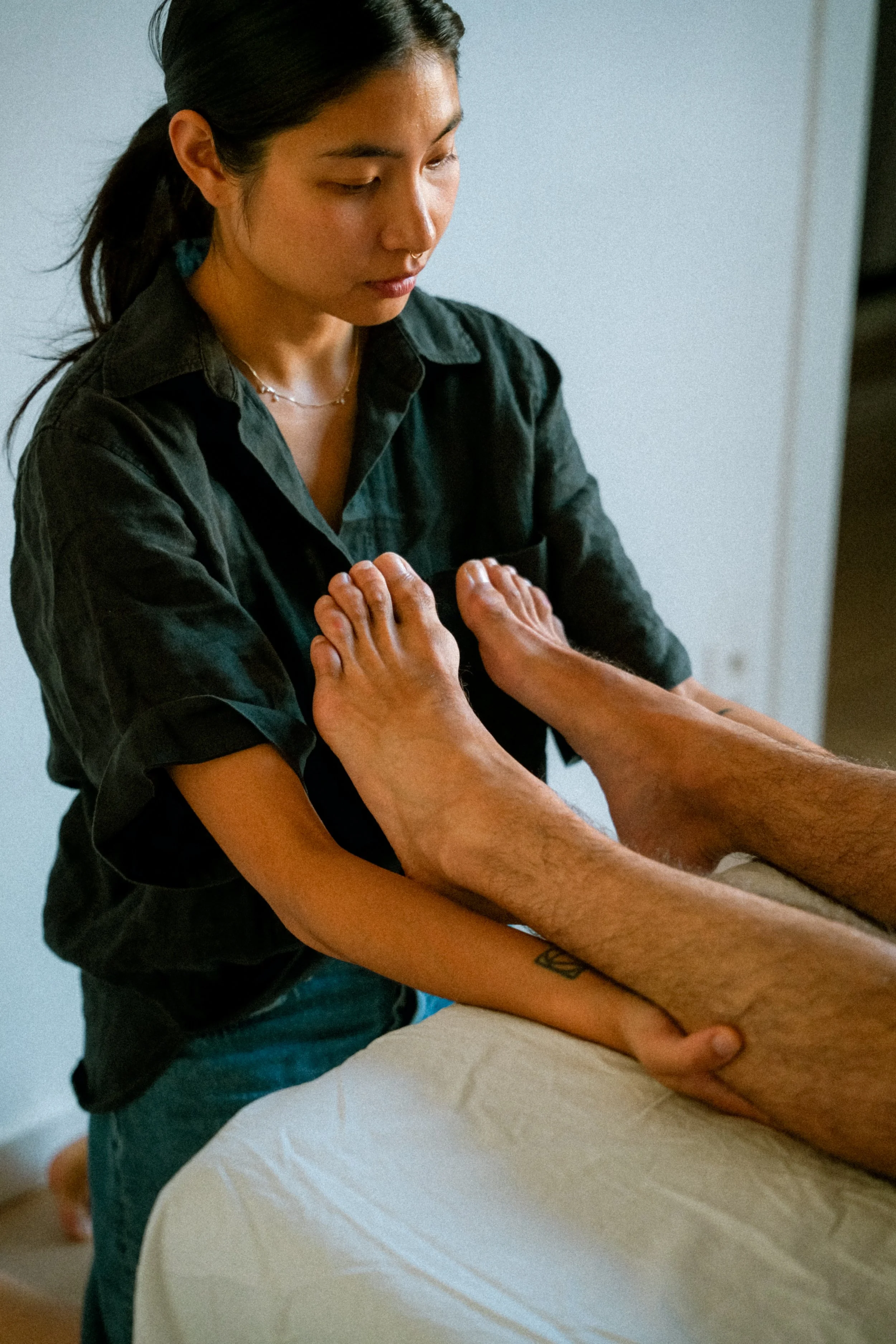 A young woman with dark hair tied back, wearing a dark shirt and layered necklaces, standing beside a person lying face down on a massage table with their arms by their sides. The young woman appears to be preparing to give a massage.