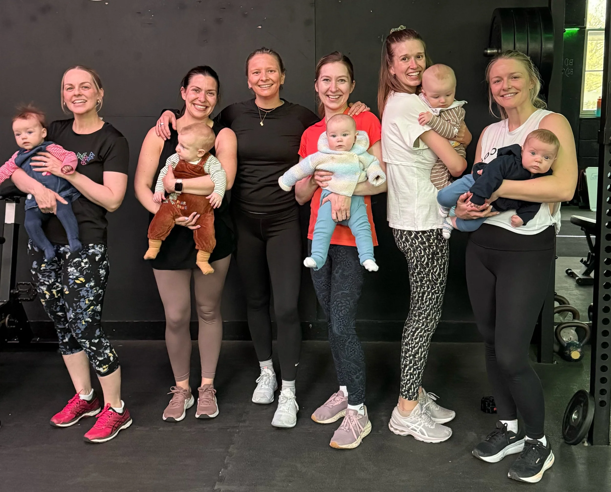 Group of seven women with their babies or young children, standing together indoors against a dark wall, smiling at the camera.