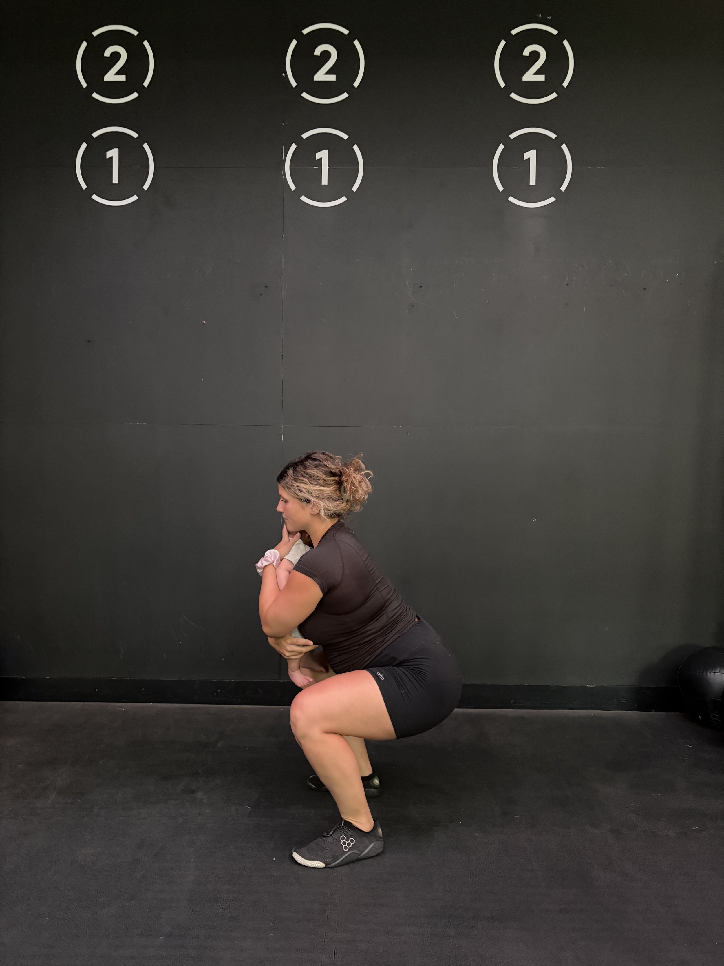 A woman in workout clothes performing a squat exercise with a person in her arms in front of a black wall.