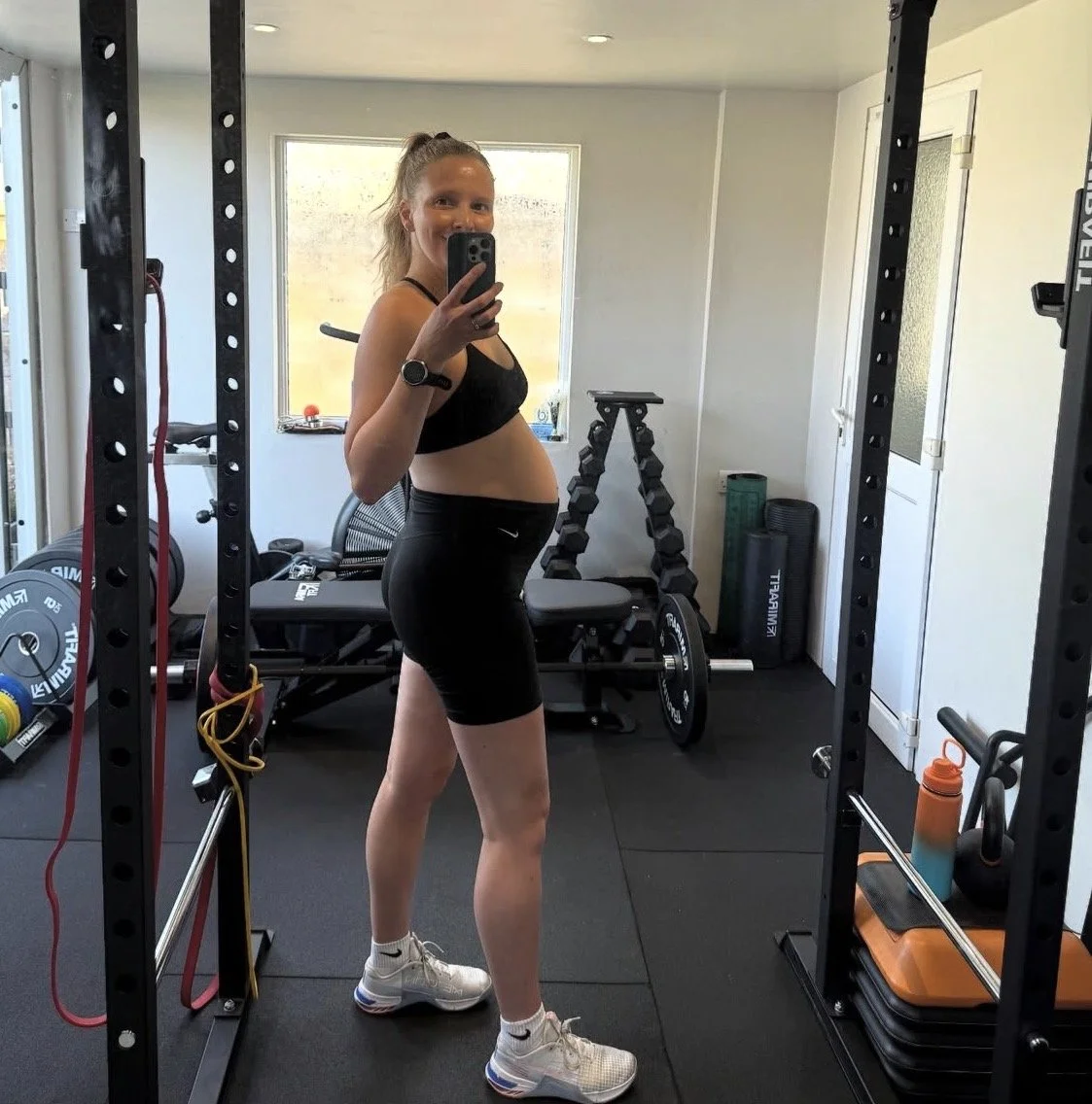 Pregnant woman taking a mirror selfie in a home gym, wearing black sports bra, black shorts, and white athletic shoes.