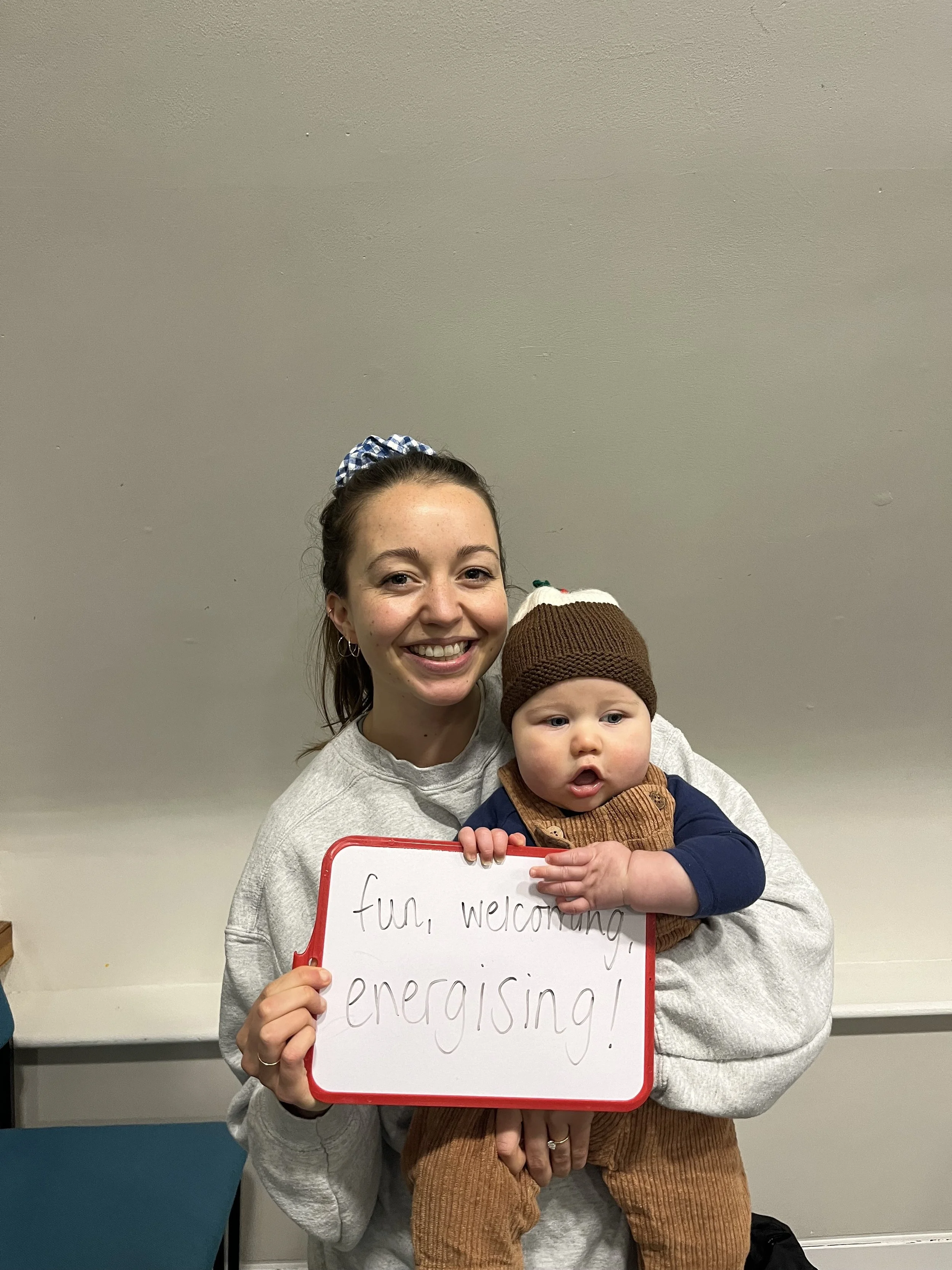 A smiling woman holding a young child, both holding a whiteboard with the words 'fun, welcoming, energising!' written on it, against a plain gray wall.