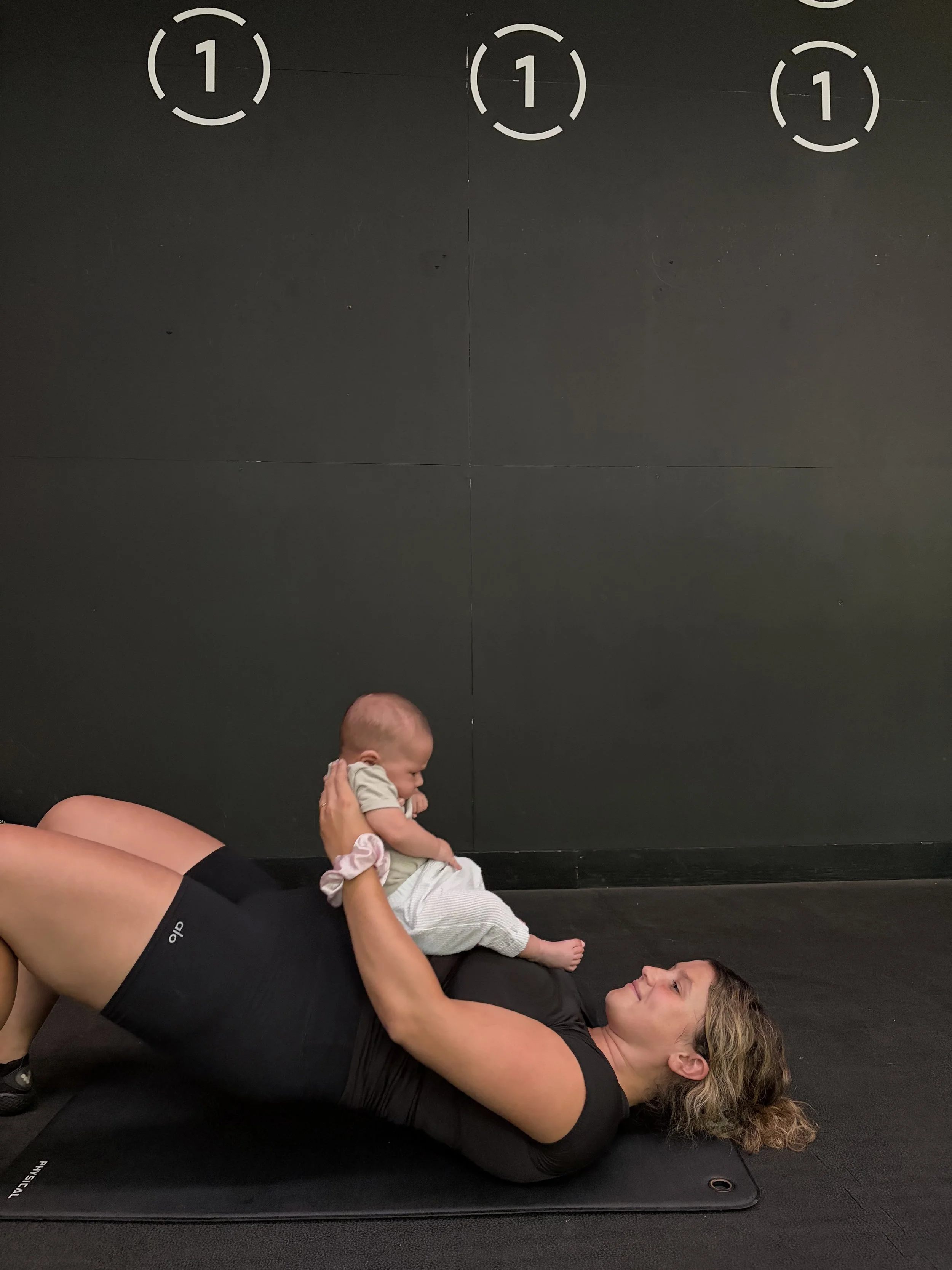 A woman lying on a black exercise mat holding a baby above her with both hands, in a black room with a black wall with white markings, and her head resting on the mat.