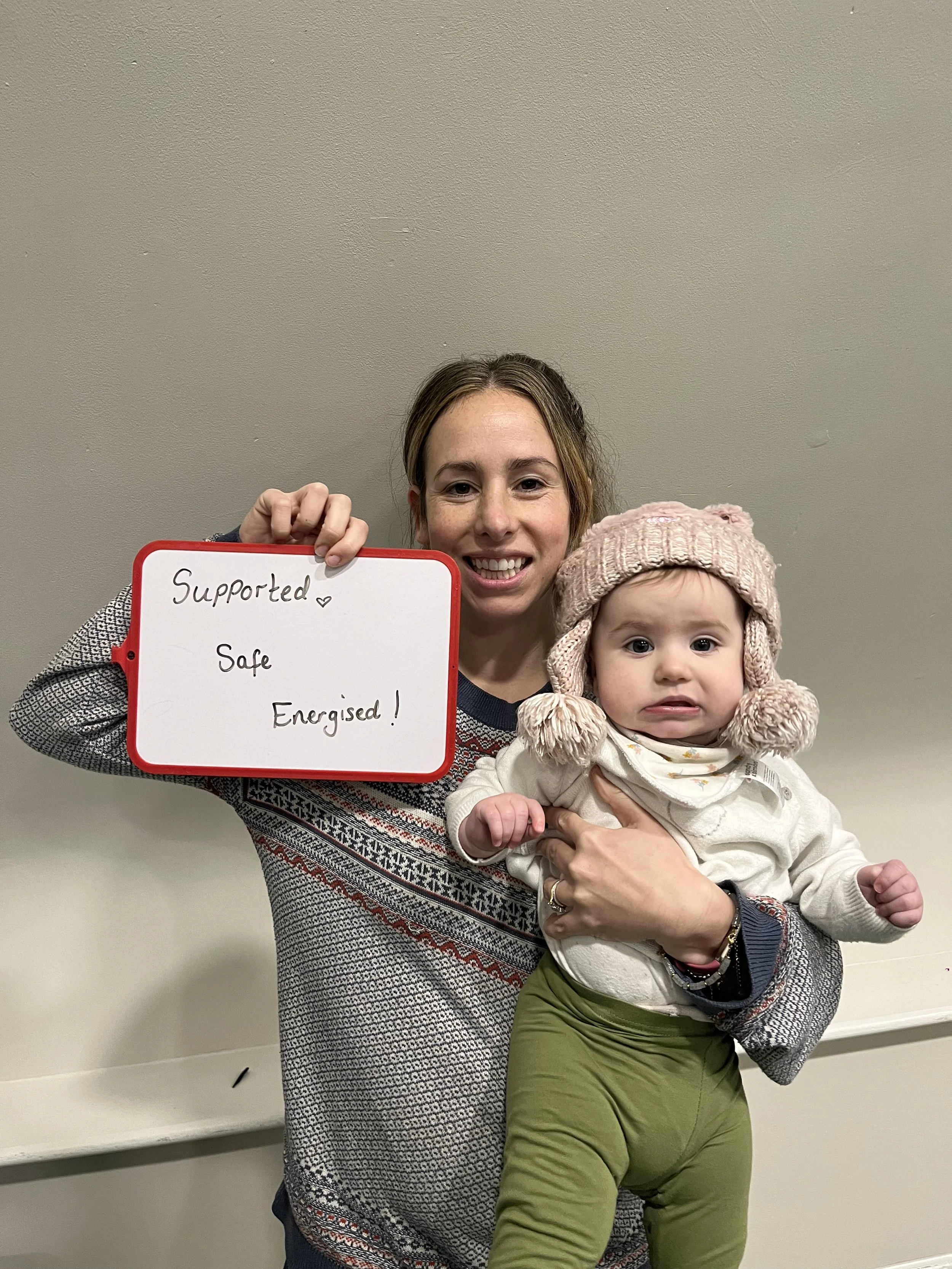 A woman holding a young child, both smiling at the camera. The woman is holding a whiteboard with the words "Supported Safe Energised!" written on it. The child is wearing a pink knit hat with earflaps and is being supported by the woman with one arm