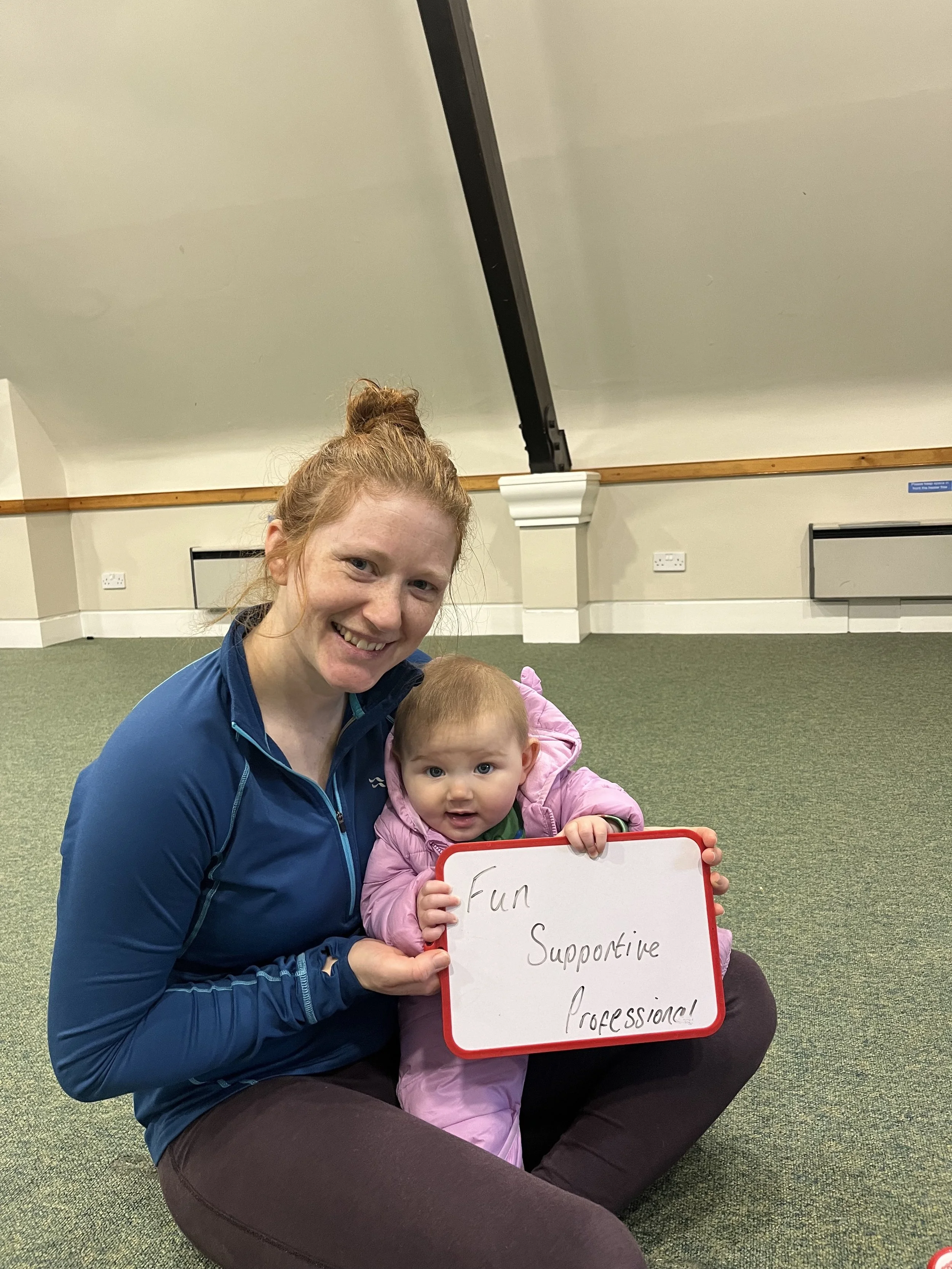 A woman and a young girl sitting on the floor in a room, holding a whiteboard that reads 'Fun, Supportive, Caring, Professional'. The woman has red hair pulled back and is smiling. The girl has light brown hair and wide eyes, also smiling.