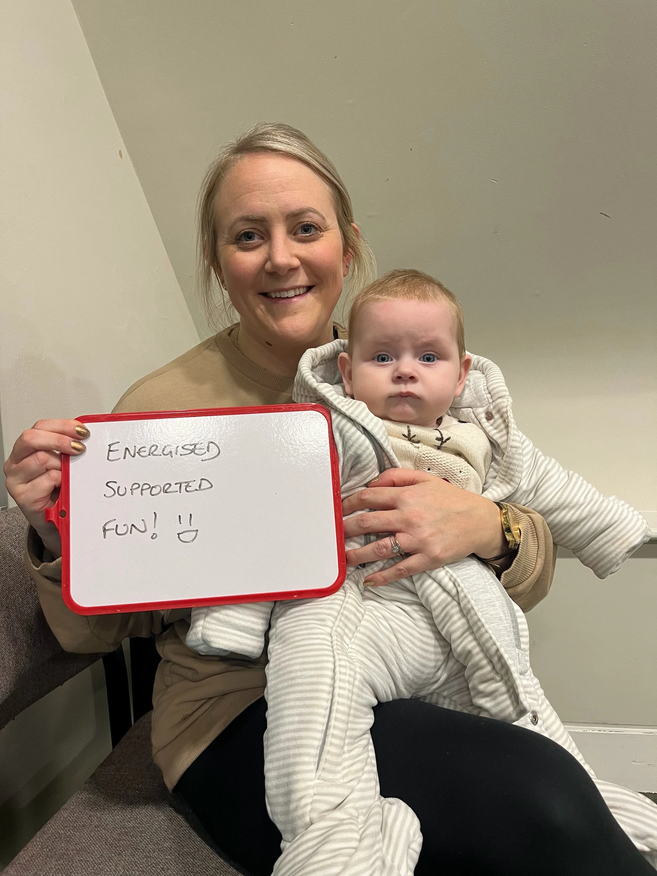 A woman smiling and holding a baby, with the baby resting on her lap, both sitting on a bench. The woman is holding a whiteboard with handwritten words 'ENERGIZED, SUPPORTED, FUN!!'.