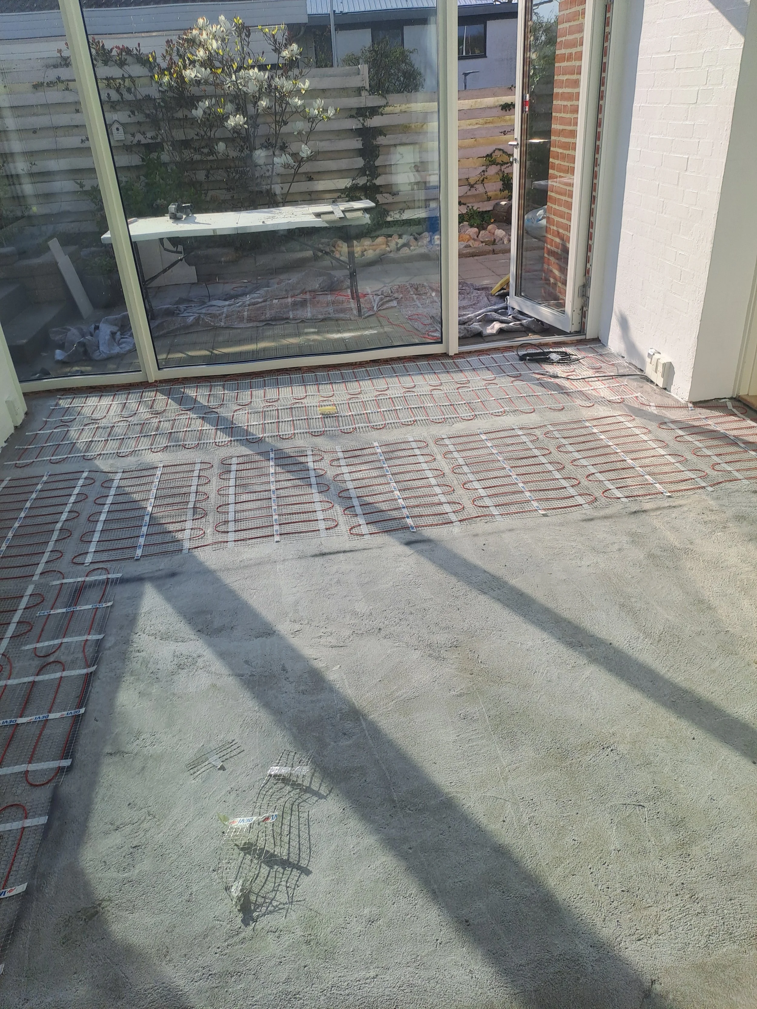 Indendørs under opførelse med varmesystem installationer synlige på gulvet, gipsvægge og en glasdør, der fører ud til en terrasse med have.