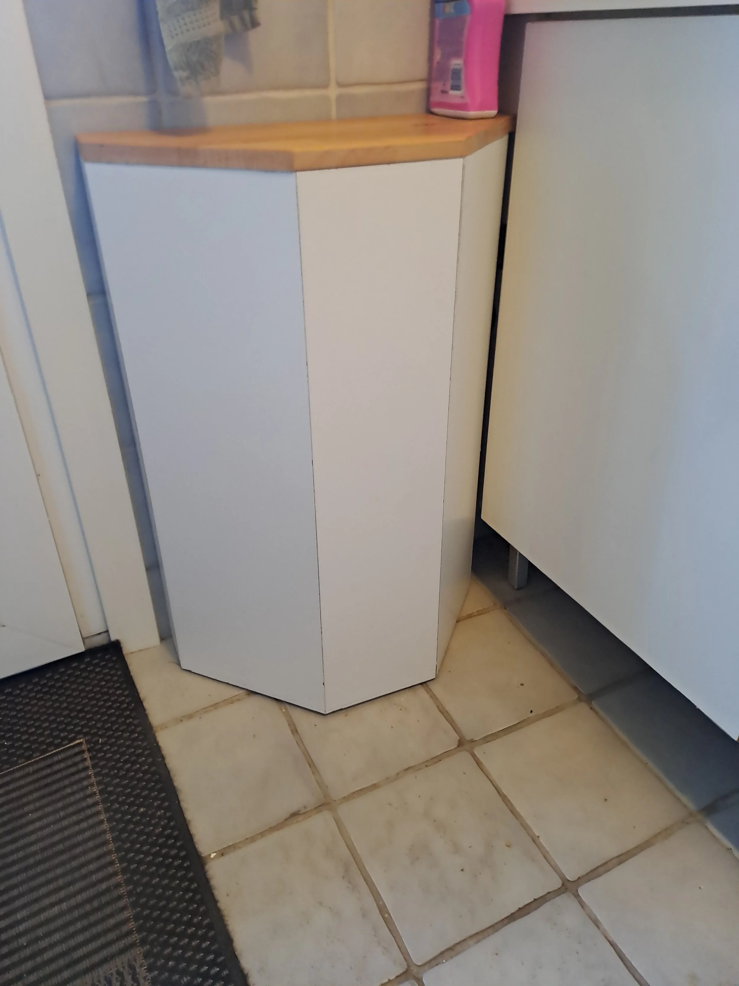 Et hjørneskab lavet af træ og hvidt materiale placeret på gulvet i et køkken. Der er en pink genstand og nogle små æsker på toppladen. Gulvet er belagt med store, beige fliser.
