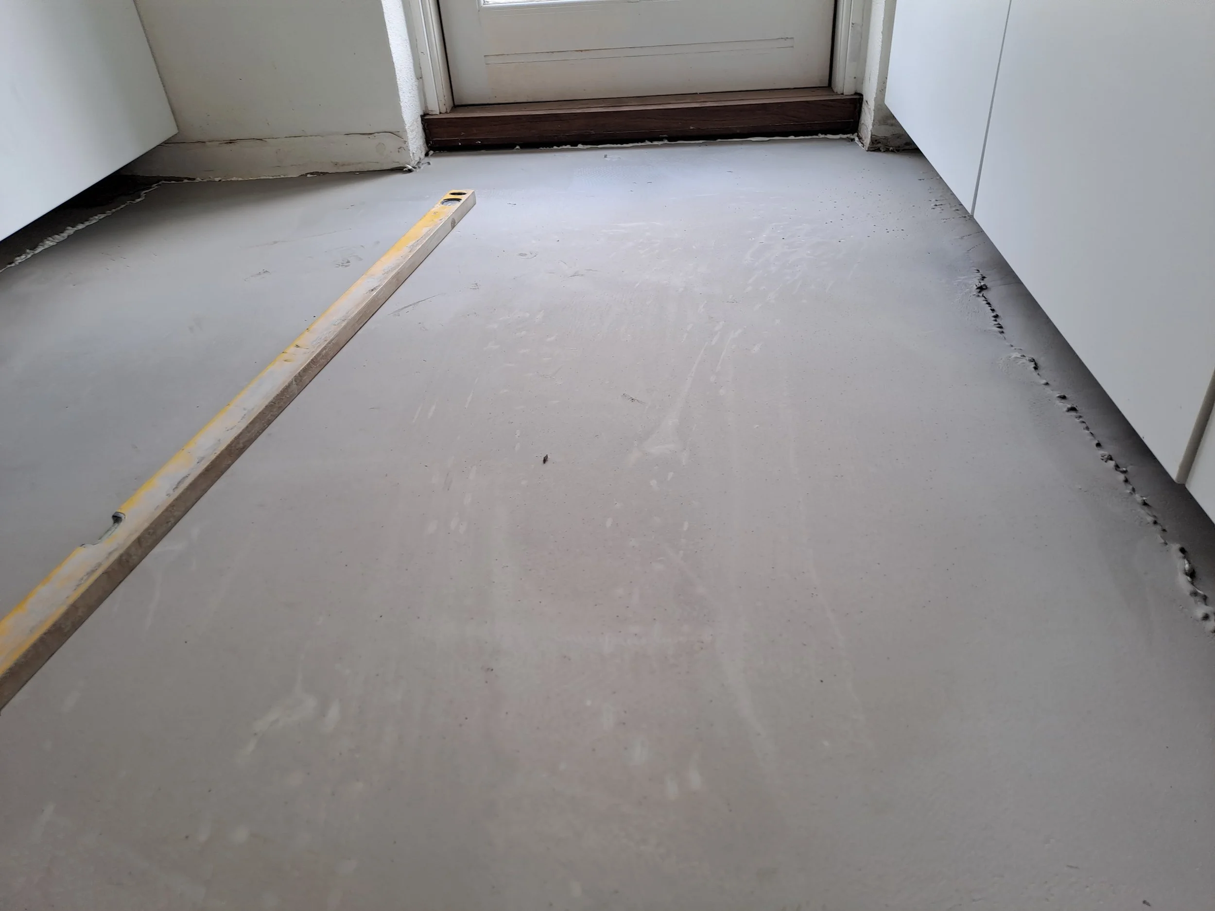 Et gulv med udgravning eller renovering, hvor en vaterpas er lagt på gulvet nær en dør ind til et rum.