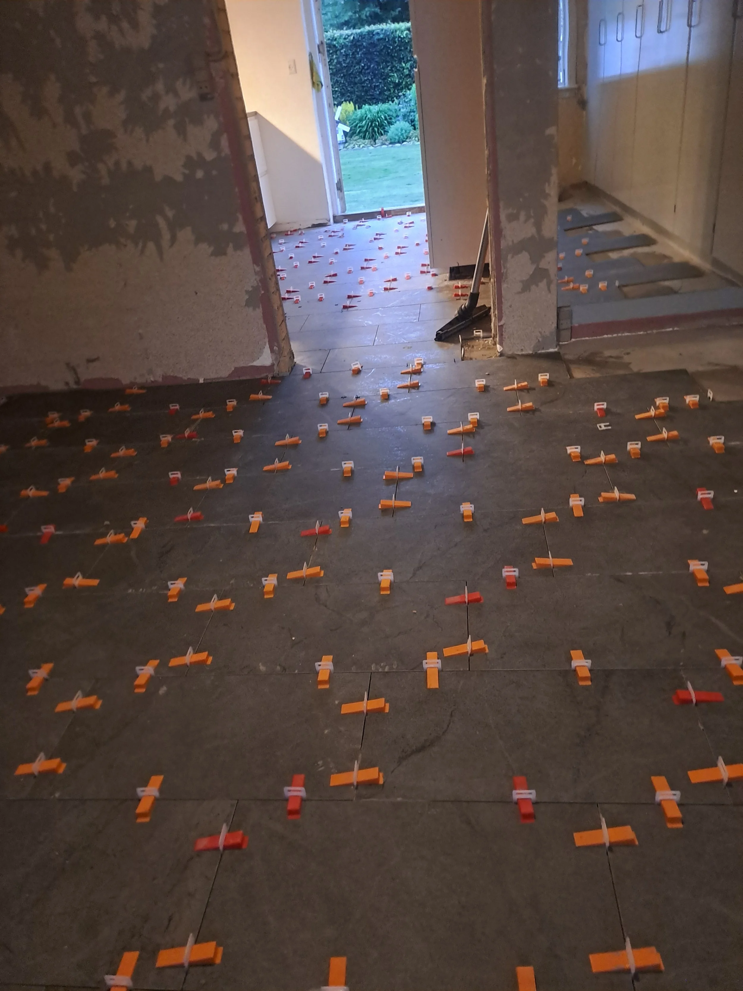 En indendørs rum under renovation med gulvfliser, hvor der er flere små orange og hvide plastik klips fastgjort på gulvet.
