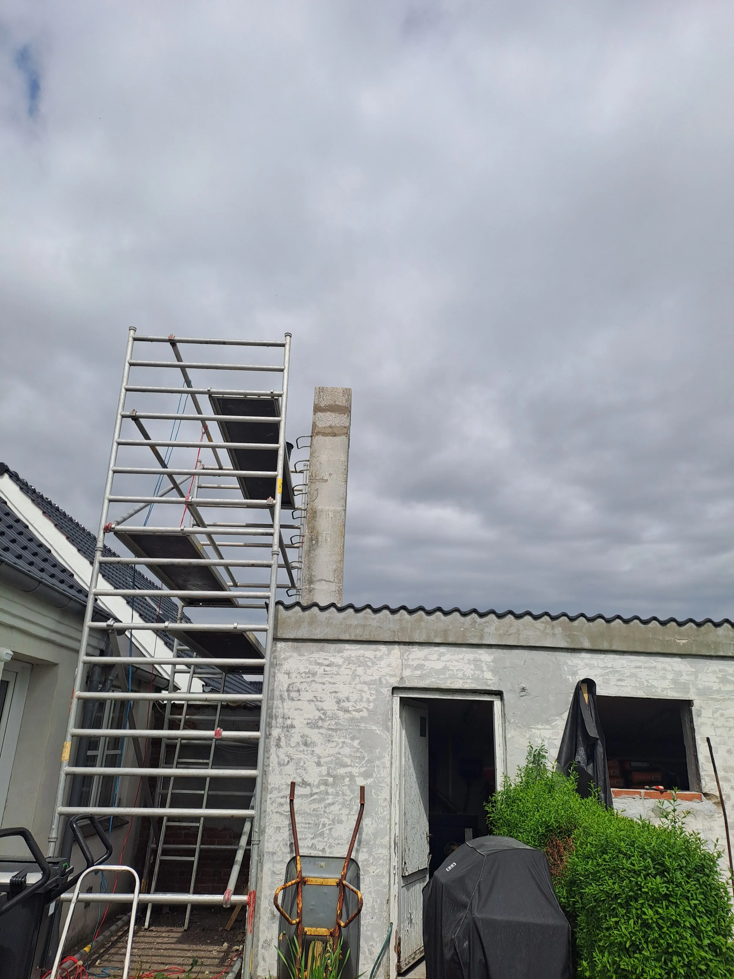 Udsigt over husets udendørs område med stillads og byggematerialer, formentlig under renovering eller vedligeholdelse, med gråt vorden og skyer på himlen.