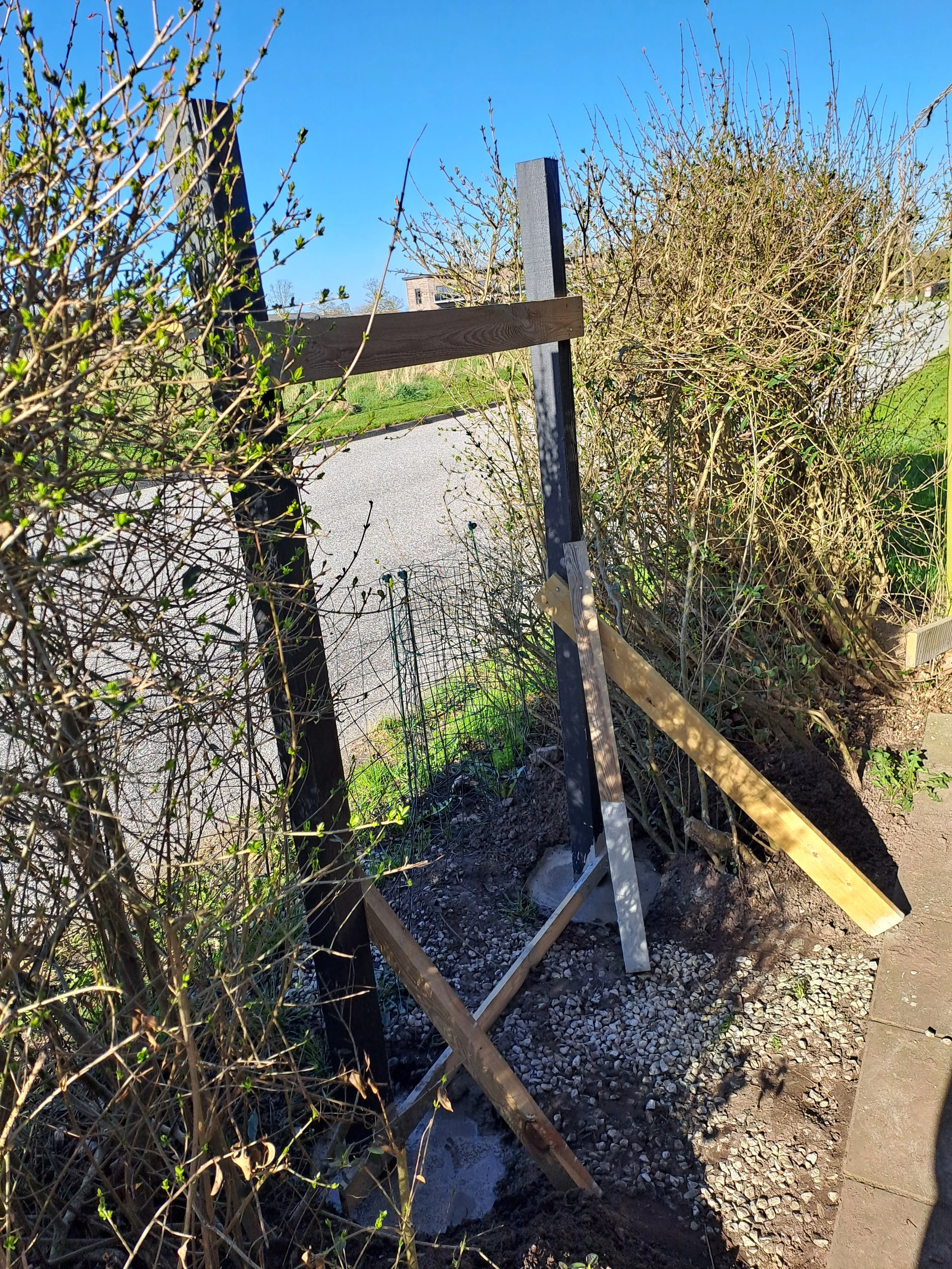 En indhegning under opførelse med træ- og metalstænger, omgivet af buske og gårdsplads, under en klar blå himmel.