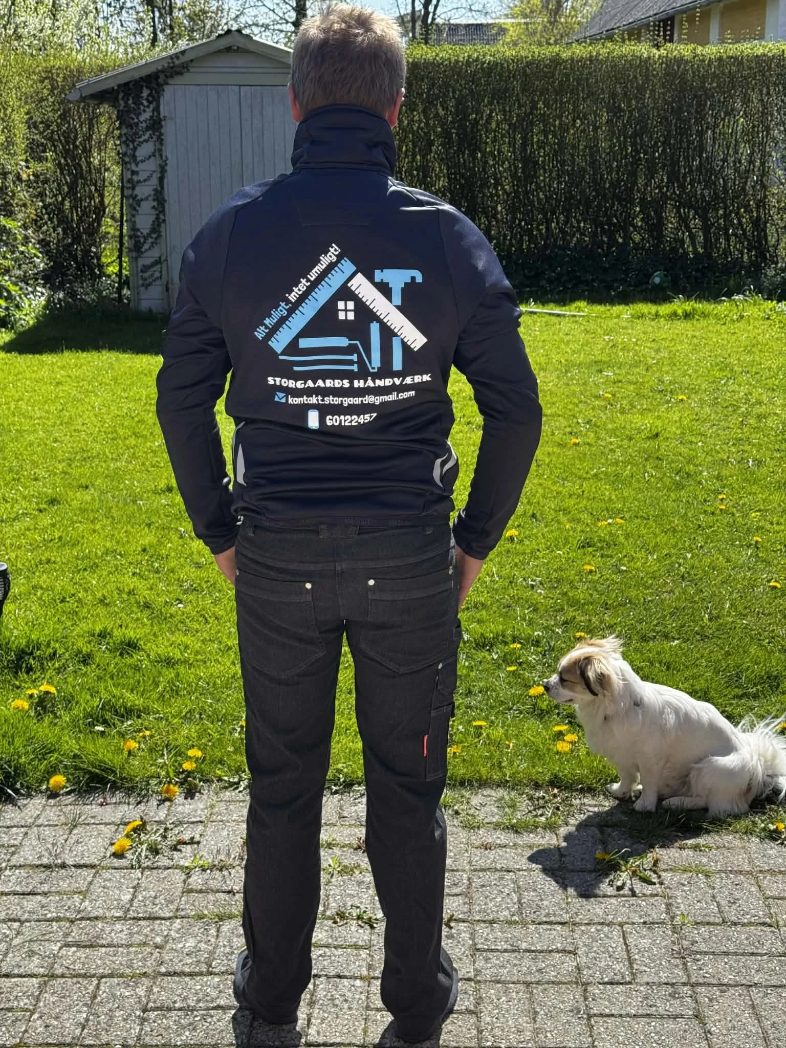En person med ryggen mod kamera står på en græsplæne med en hund til højre, både personens jakke og hunden vender mod en hæk. Personens jakke har en logo og kontaktinformation på ryggen.