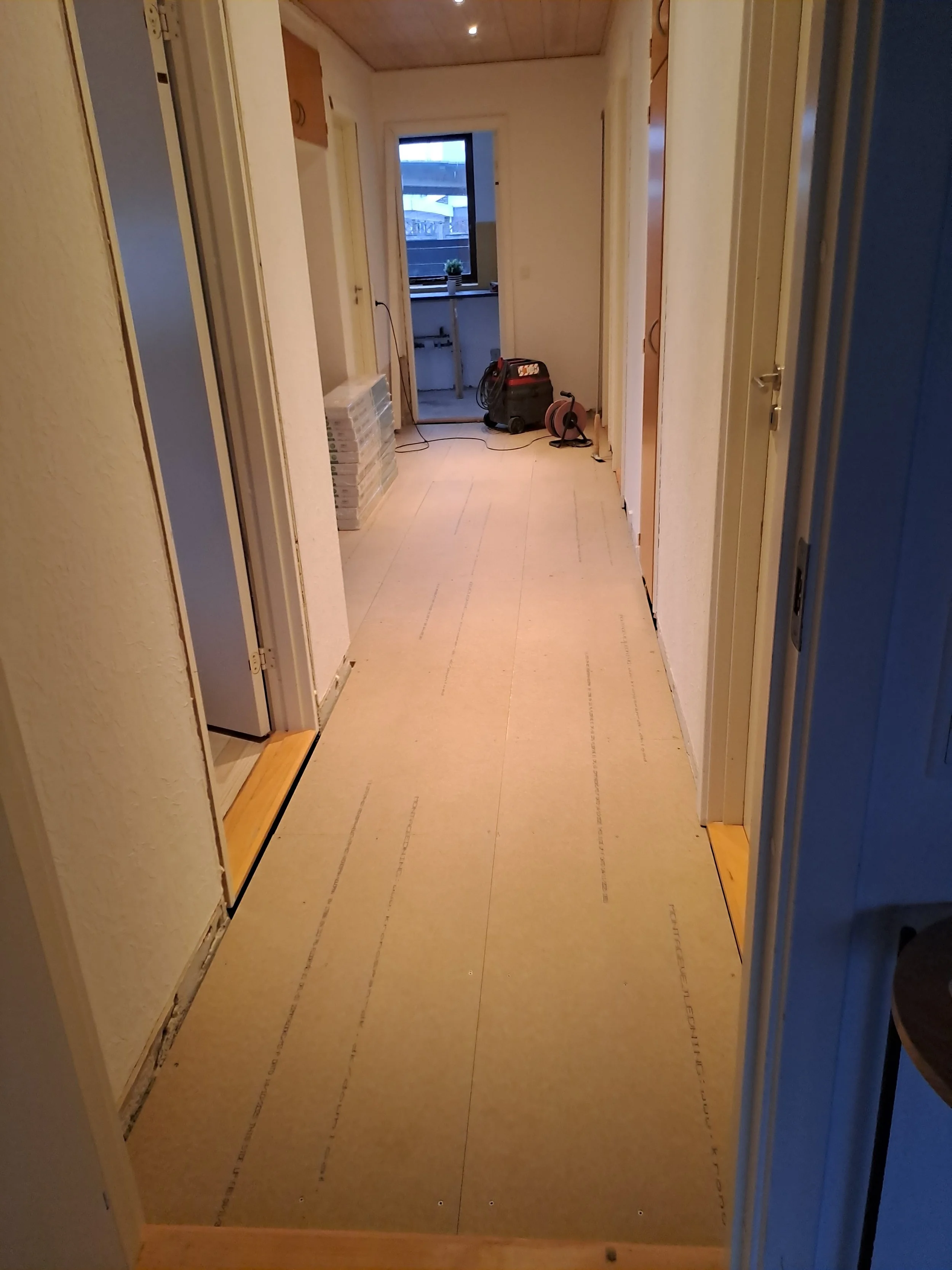 Indretningsarbejde i gang med nymalet væg og gulvbeskyttelse. Der er værktøj, såsom kabelrulle og sprøjte, samt nogle byggematerialer i rummet.
