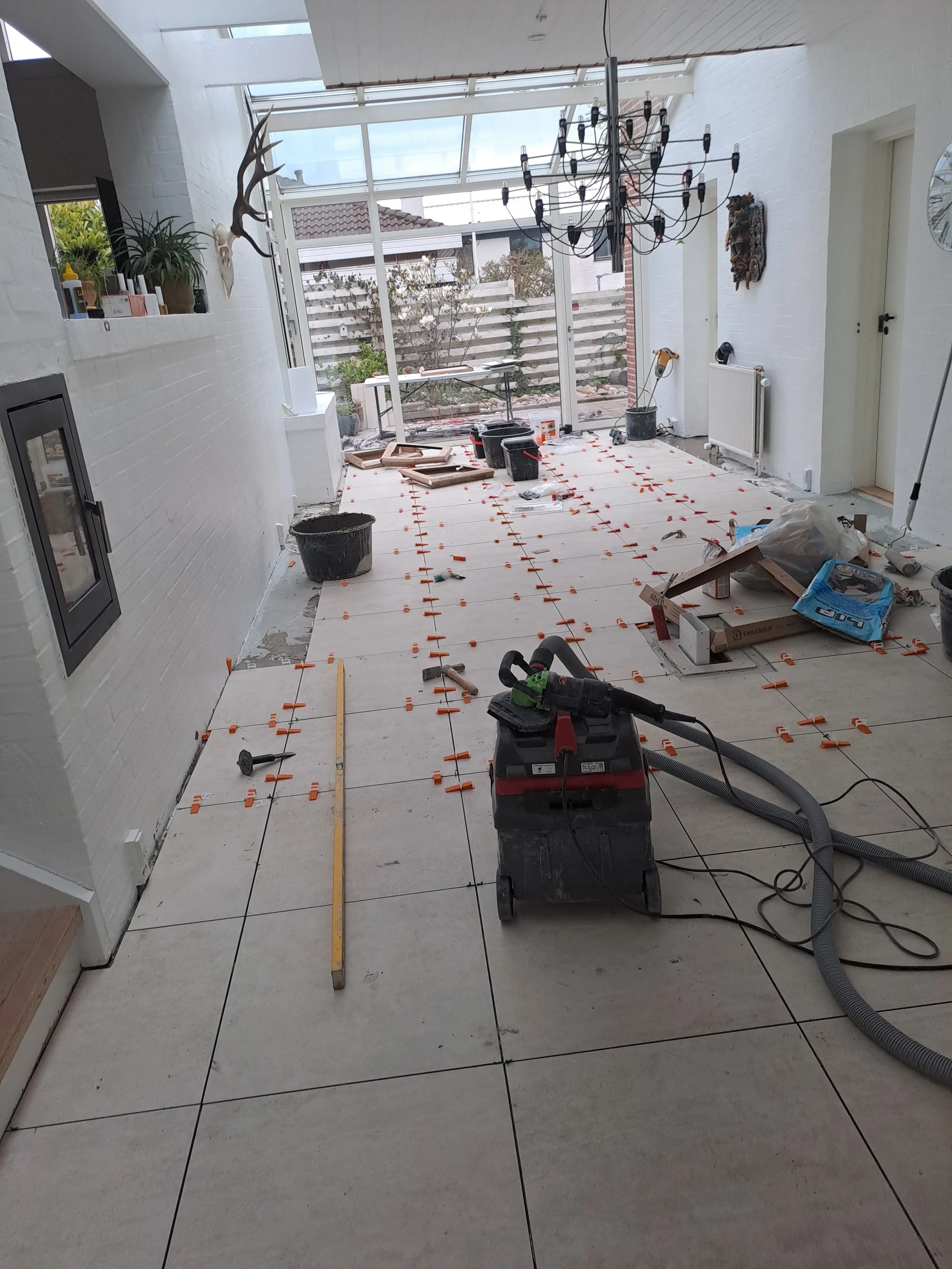 Indendørs rum under renovering med gulvfliser, værktøj, spand og byggeudstyr. Der er et stort vindue, der vender ud mod en have med planter og en træ-dæk. Der er også et smykke eller metalkunstværk på væggen.