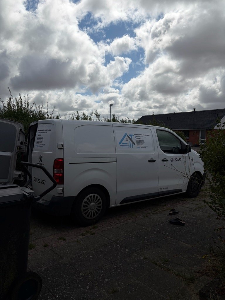 En hvid varevogn parked lunt mod en hus, med en logo og kontaktoplysninger på siden, under en skyet himmel.
