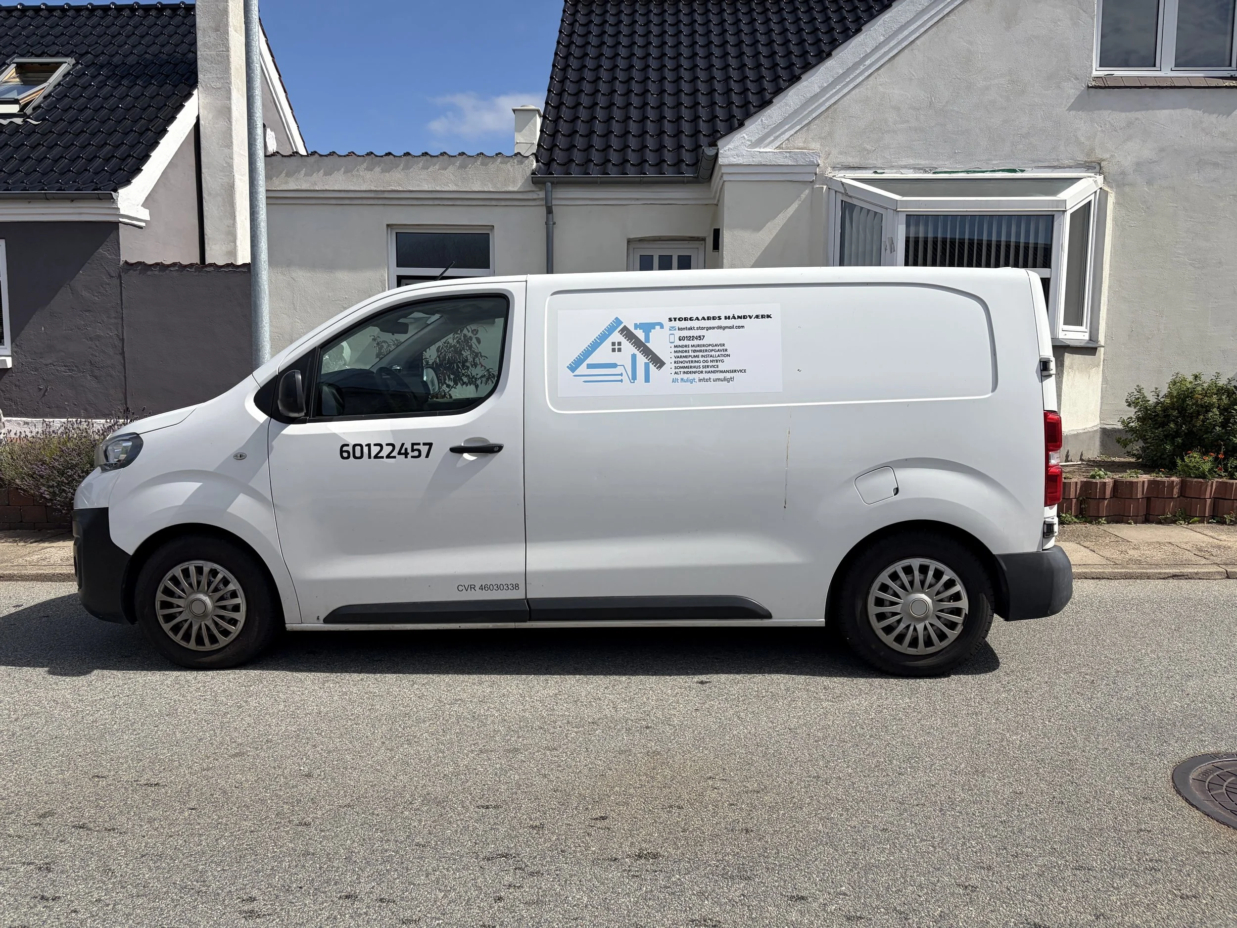 En hvid firmabil med kontaktoplysninger for Storgårds Håndværk, parkeret på en vej foran huse med sorte og hvide tage.