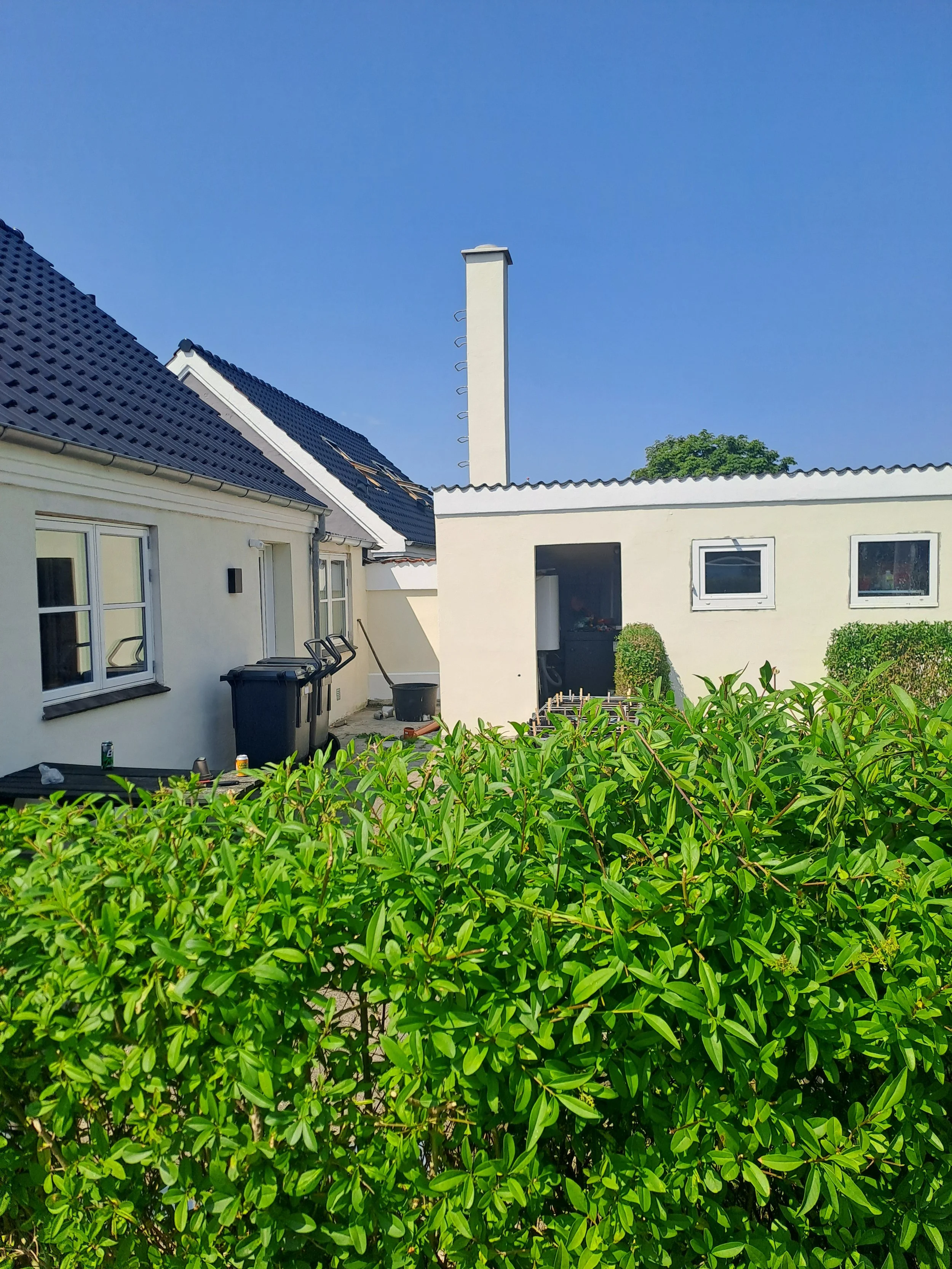 Hus med hvide vægge og blå tagplader, små vinduer, en skorsten, grønne buske og en klar blå himmel.